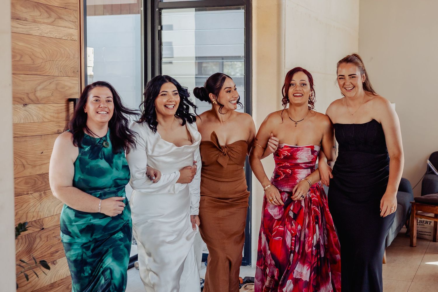 Five women in formal dresses laughing together, including a bride in white satin gown with bridesmaids in colorful attire.