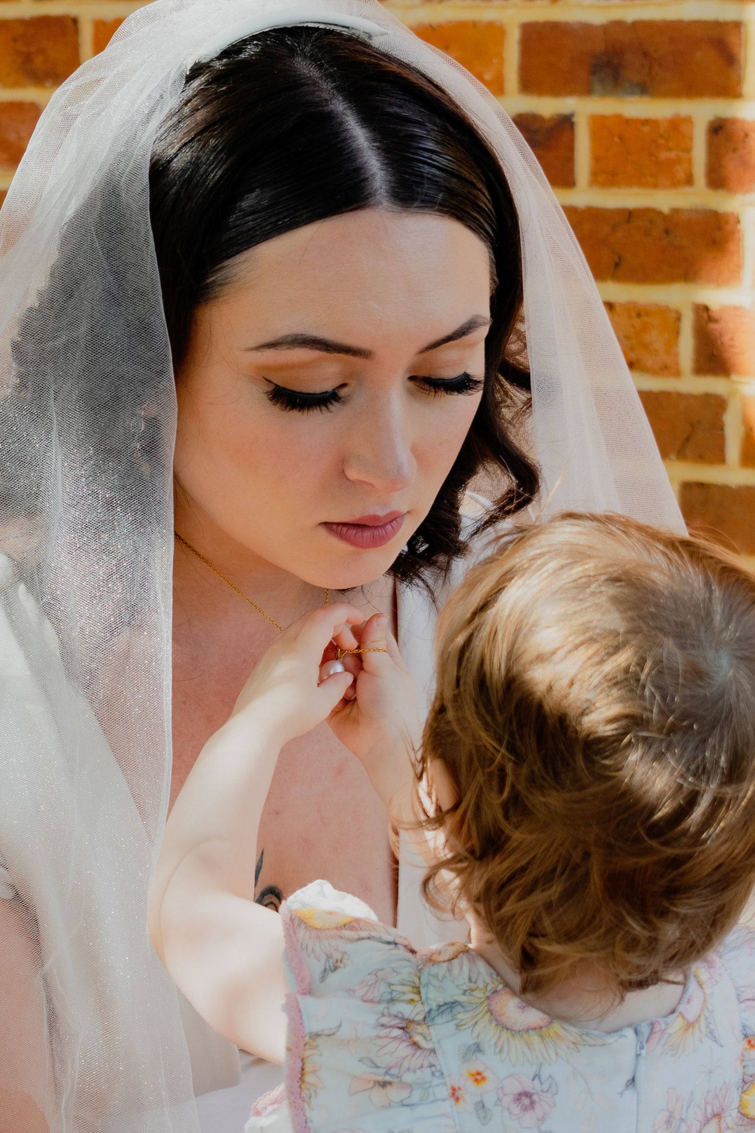 Sweet intimate moment between bride in white veil and small child against brick wall background.