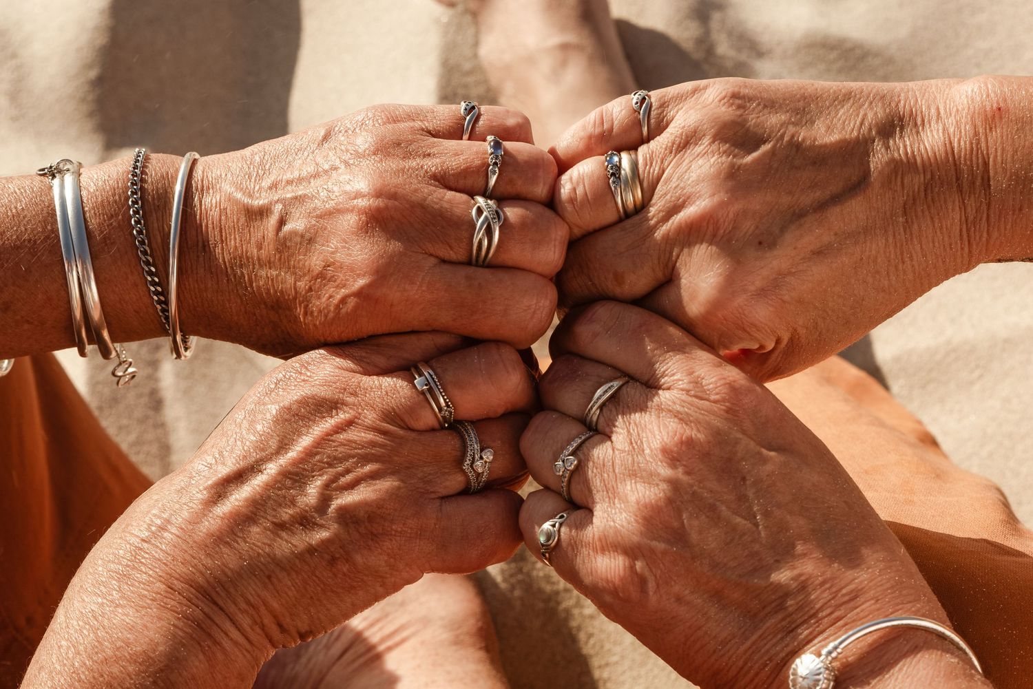 Detail shot of hands with jewelry and rings clasped together on sandy backdrop.