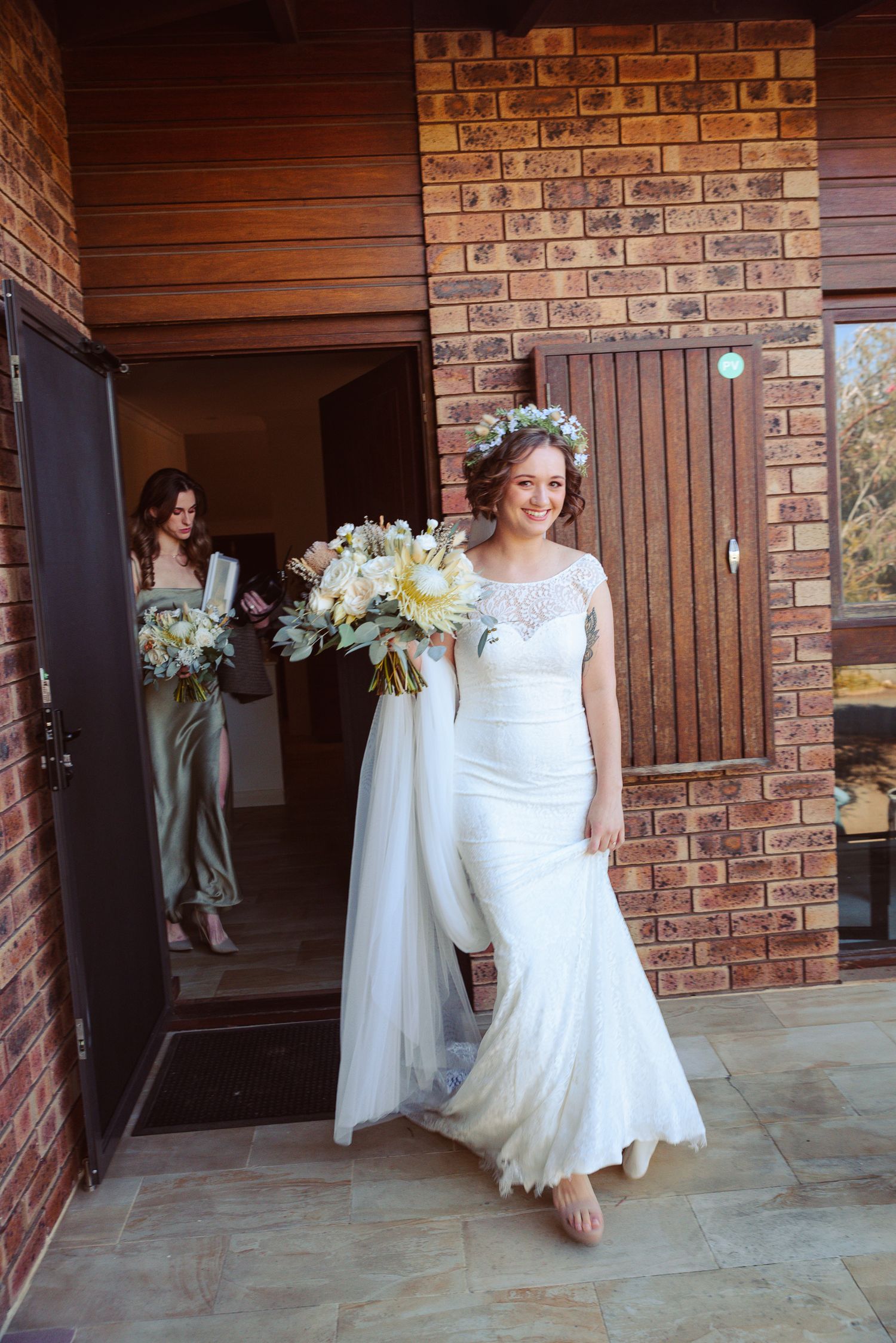Smiling bride in white gown and floral crown holds bouquet, stepping out of brick building with bridesmaid behind her.