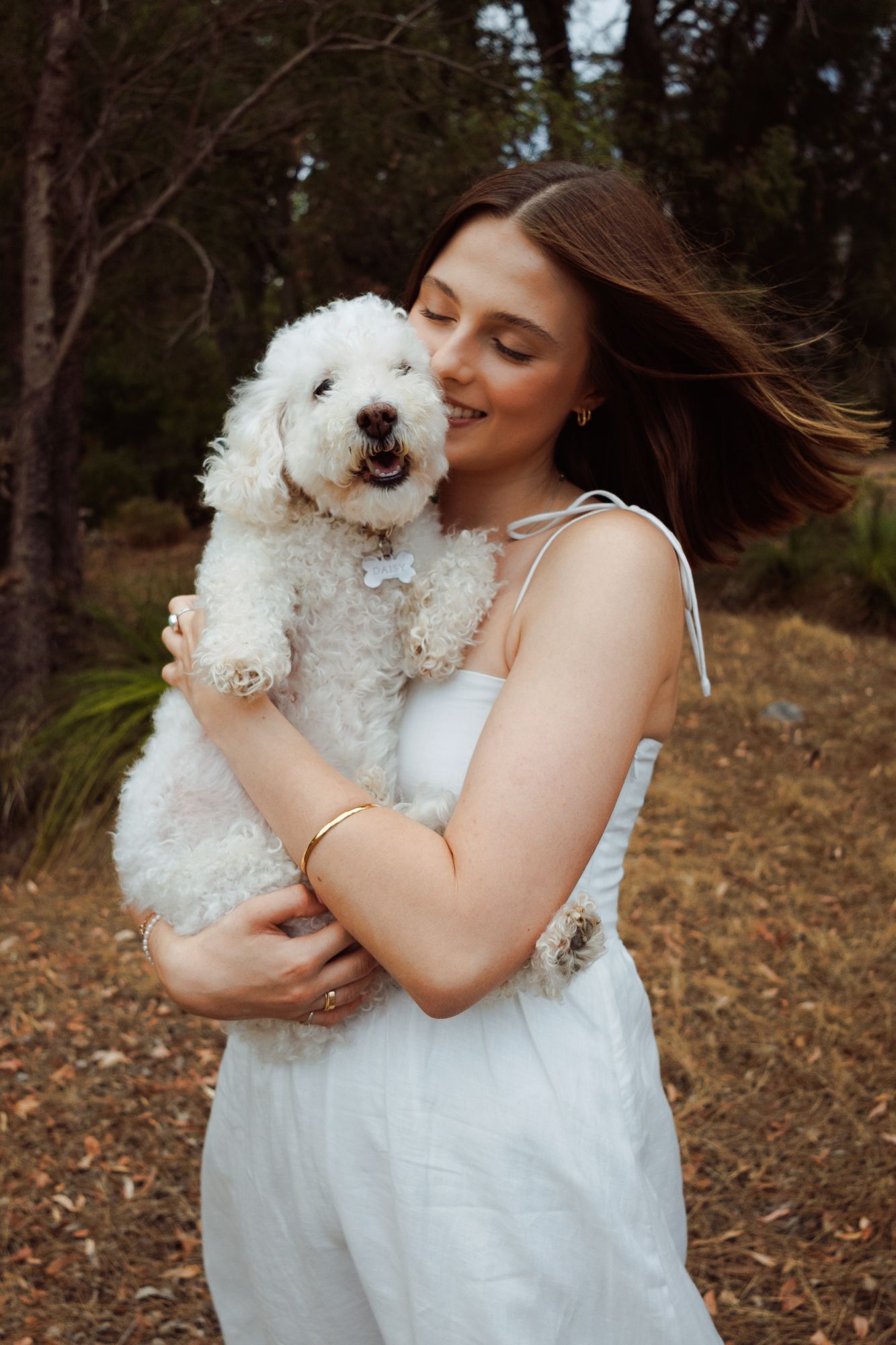Adorable white fluffy dog being cuddled and kissed by someone in a white dress in an outdoor setting.