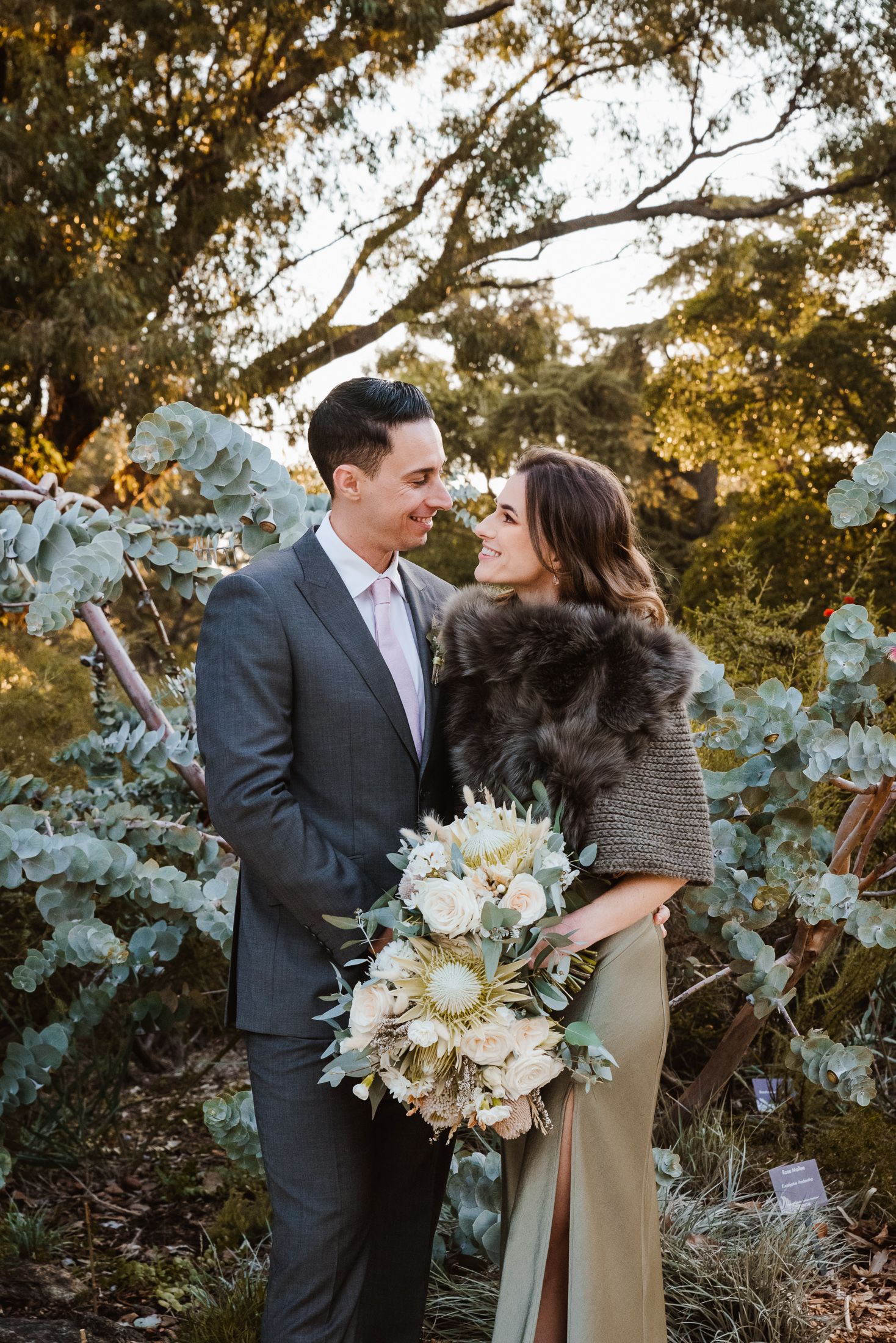 A winter wedding photoshoot with a couple embracing amidst eucalyptus trees and holding a white bouquet.