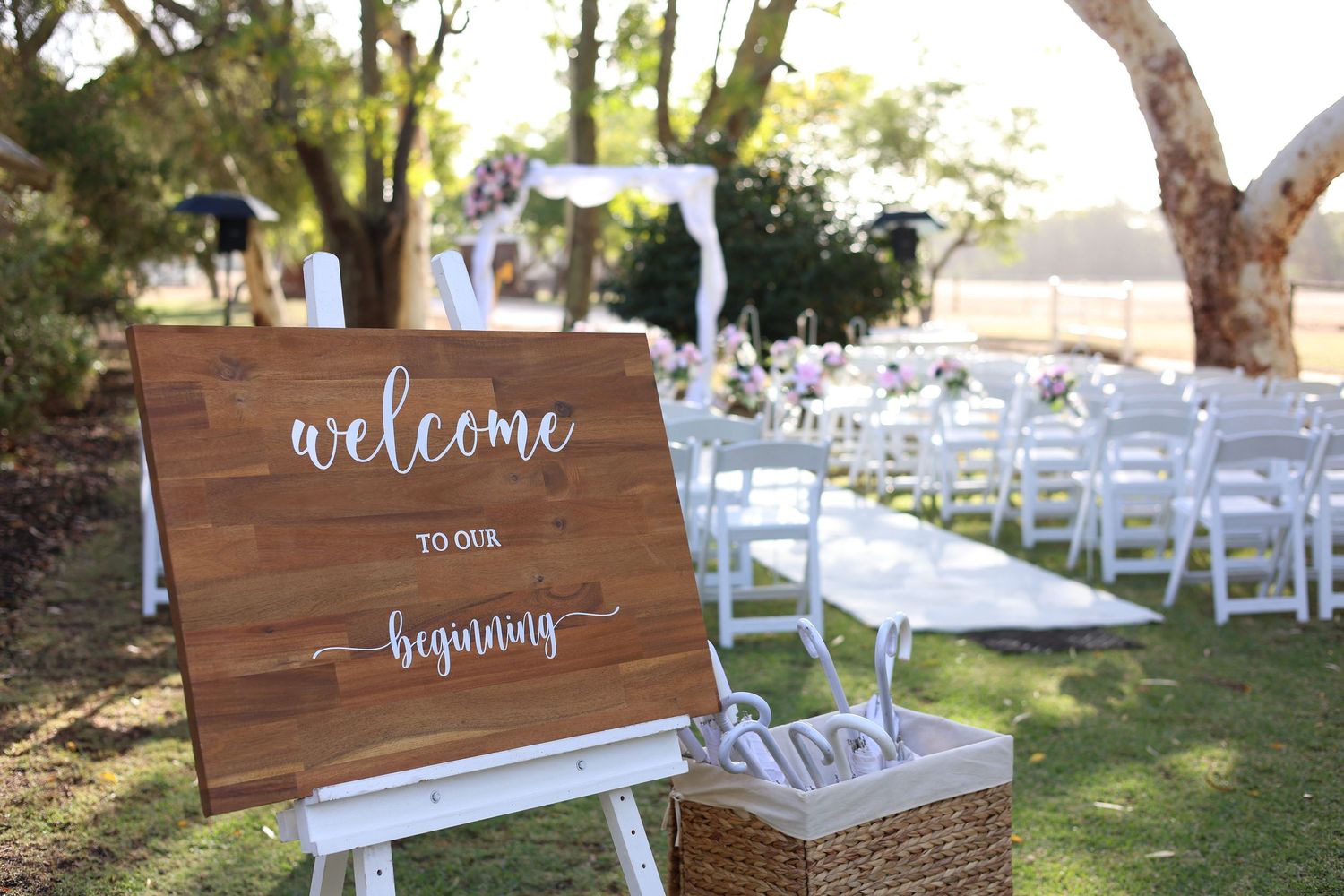 Rustic wooden welcome signs and white chairs set up for an outdoor wedding ceremony in a garden setting.