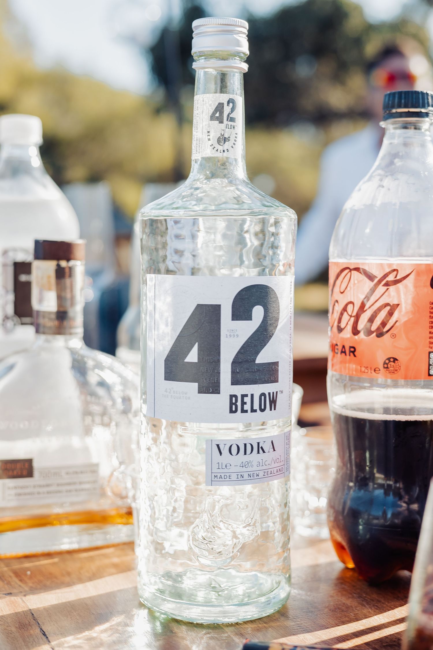 42 Below New Zealand vodka bottle displayed outdoors at a bar setup alongside Coca-Cola and other spirits.
