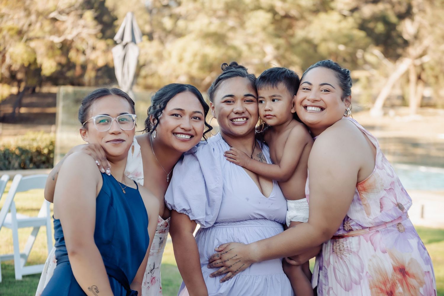 Five women smiling and posing together outdoors at a garden event, one holding a young child, with trees and chairs in background.