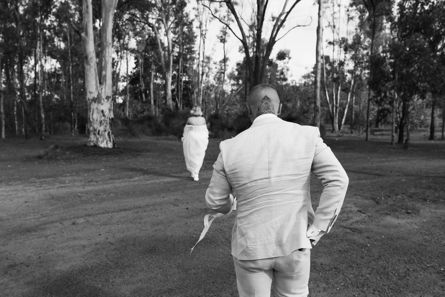 Man in white suit holding a white cockatoo bird, standing on a dirt path surrounded by eucalyptus trees.