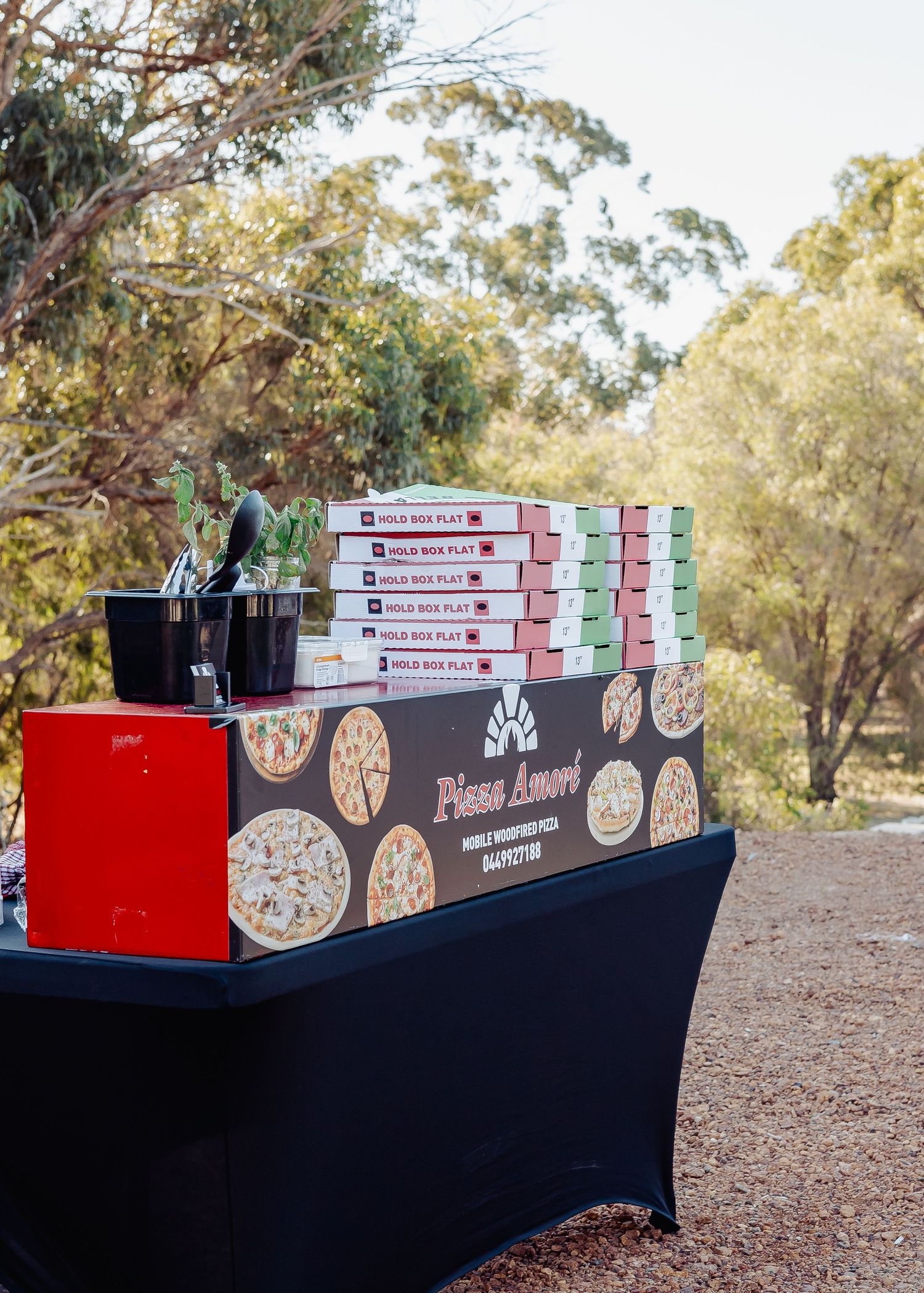 Stack of Pizza Amore pizza boxes displayed on a black table outdoors surrounded by trees at a catering event.