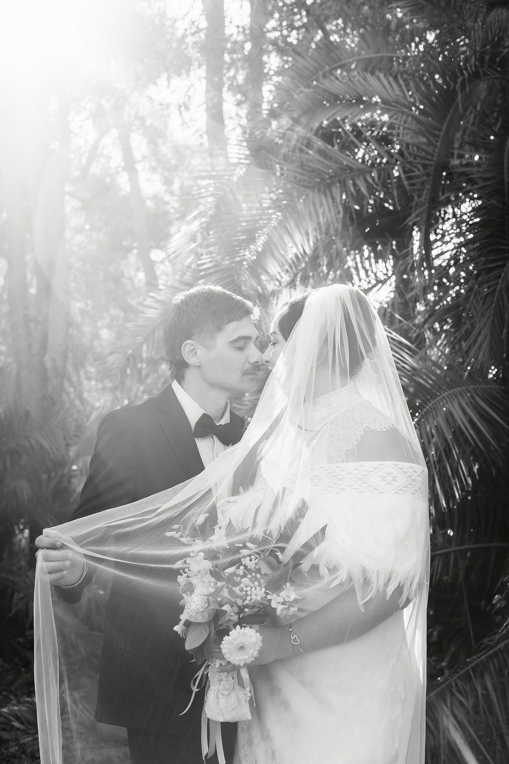 Black and white photo of a bride and groom sharing a kiss under a flowing veil, surrounded by lush tropical palms.