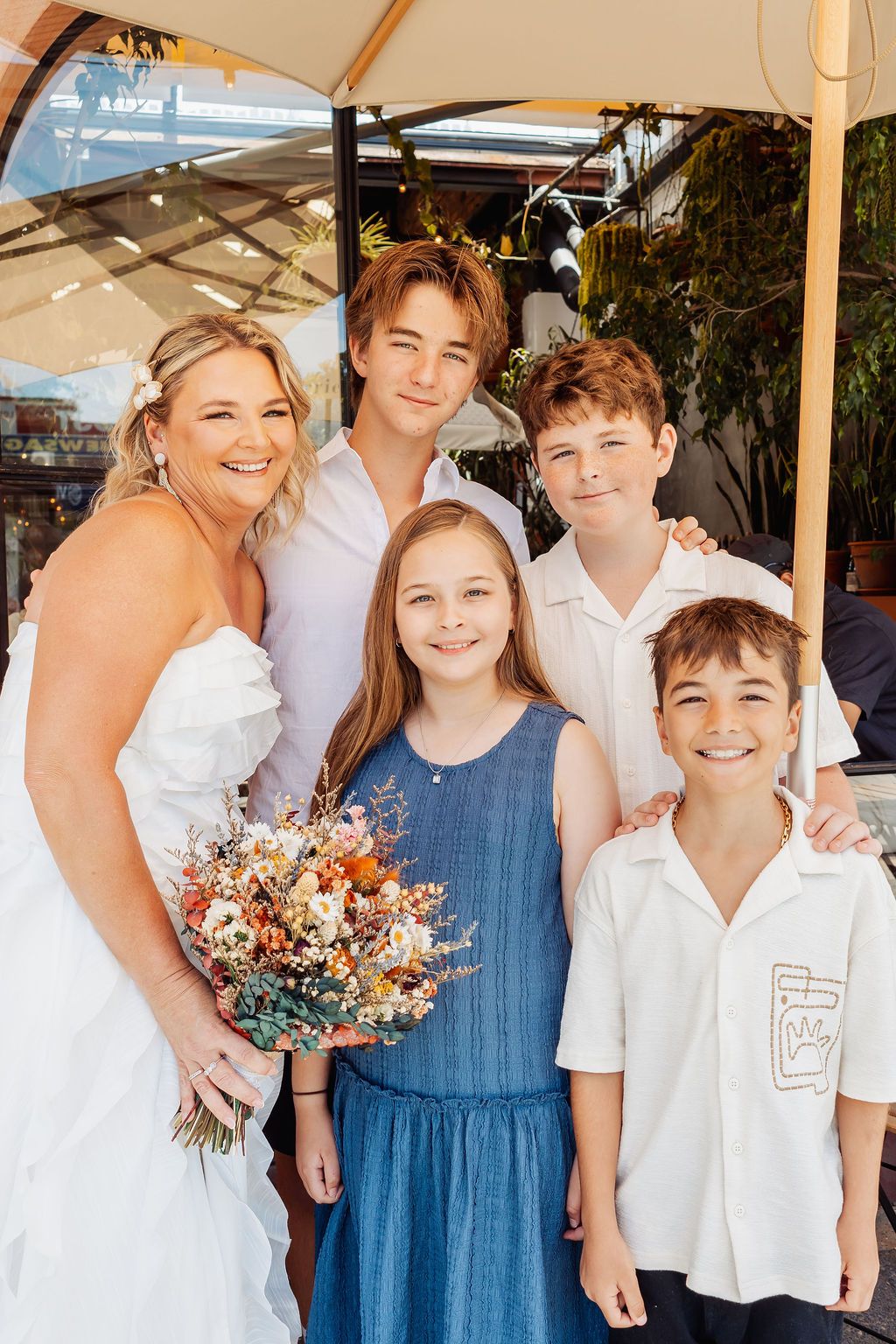 Bride in white dress holds colorful bouquet posing with four children at an outdoor wedding venue.