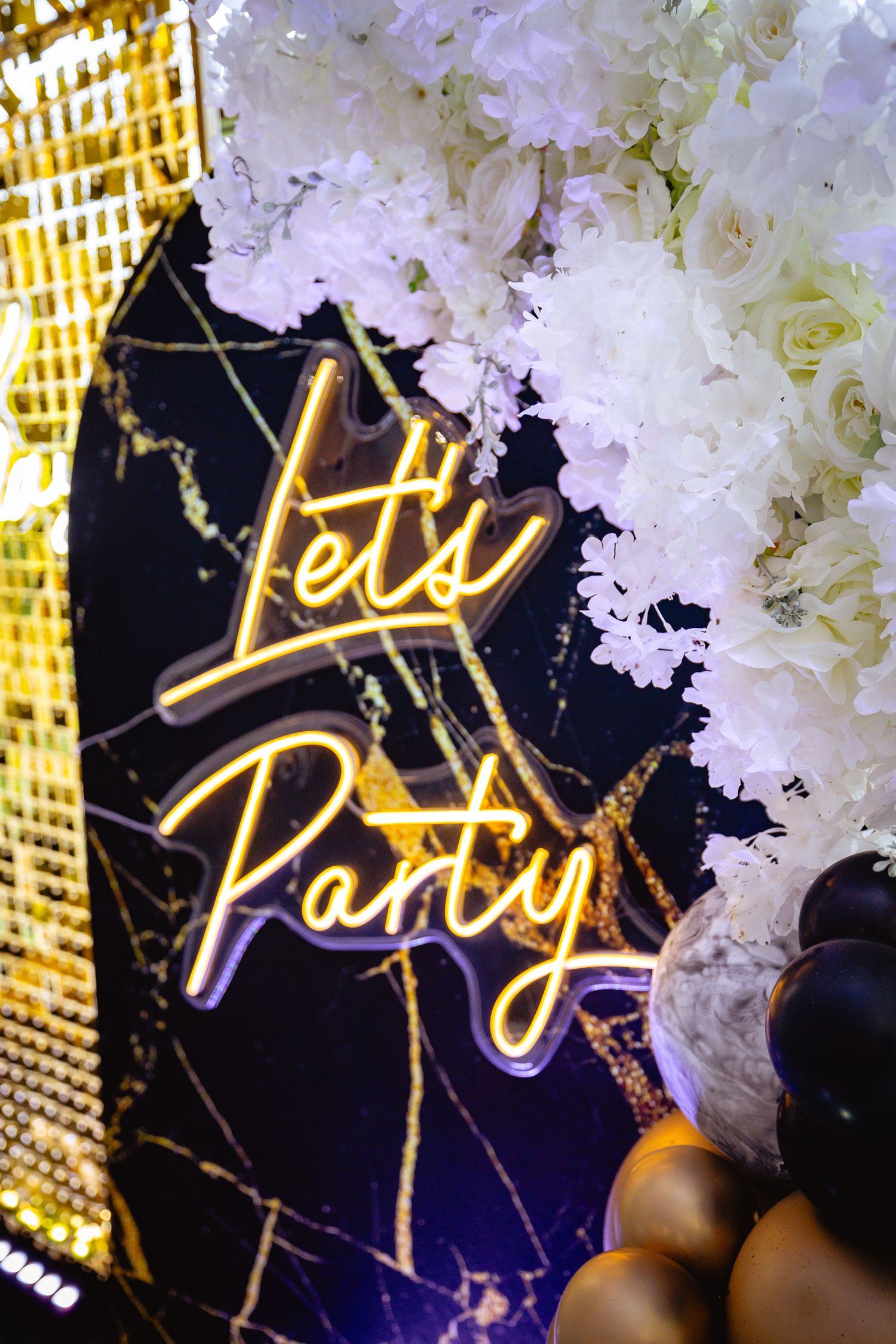 Elegant gold 'Lets Party' neon sign display with white floral decorations against dark background.