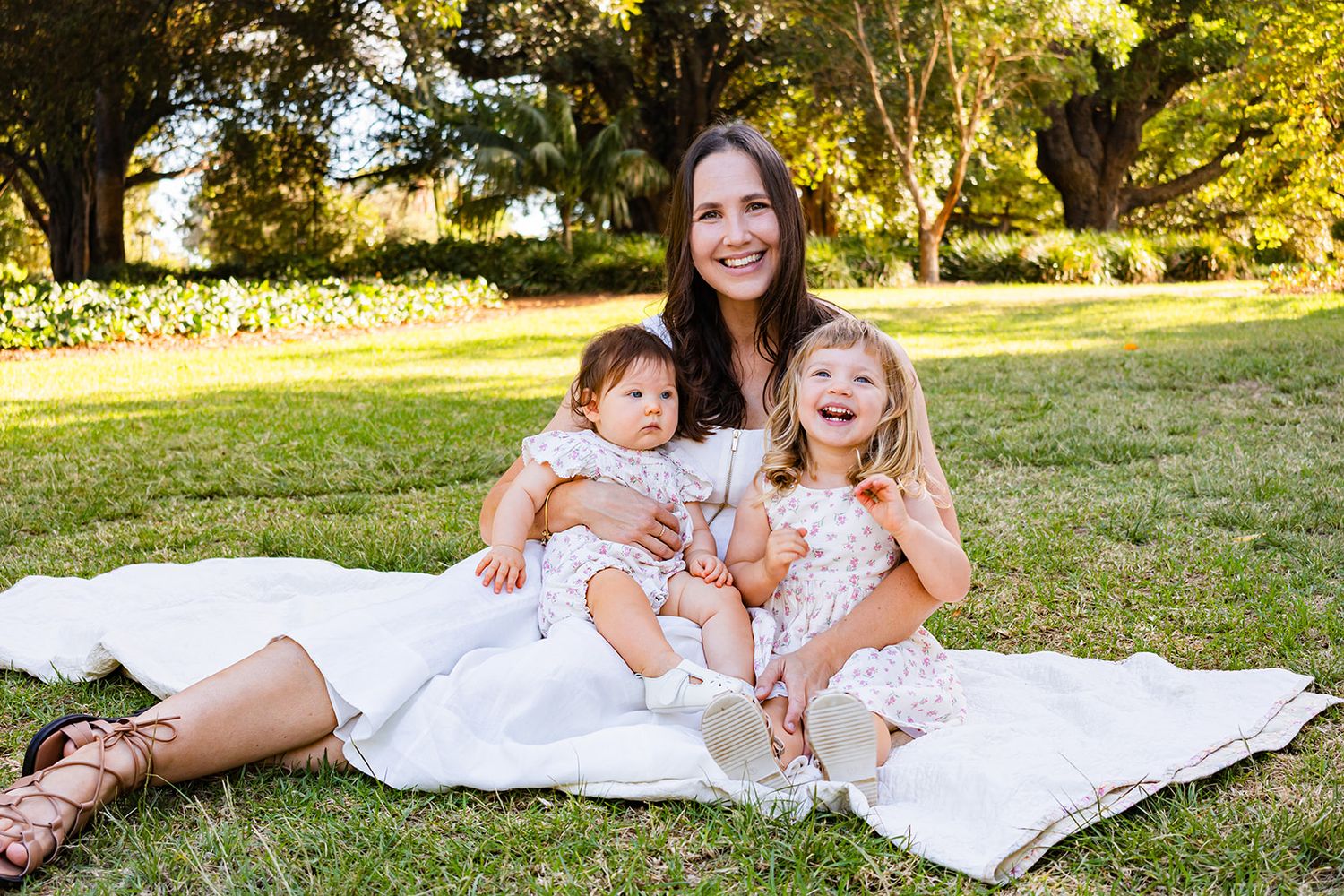Family enjoying an outdoor photoshoot together on a white blanket in a sunny park setting.