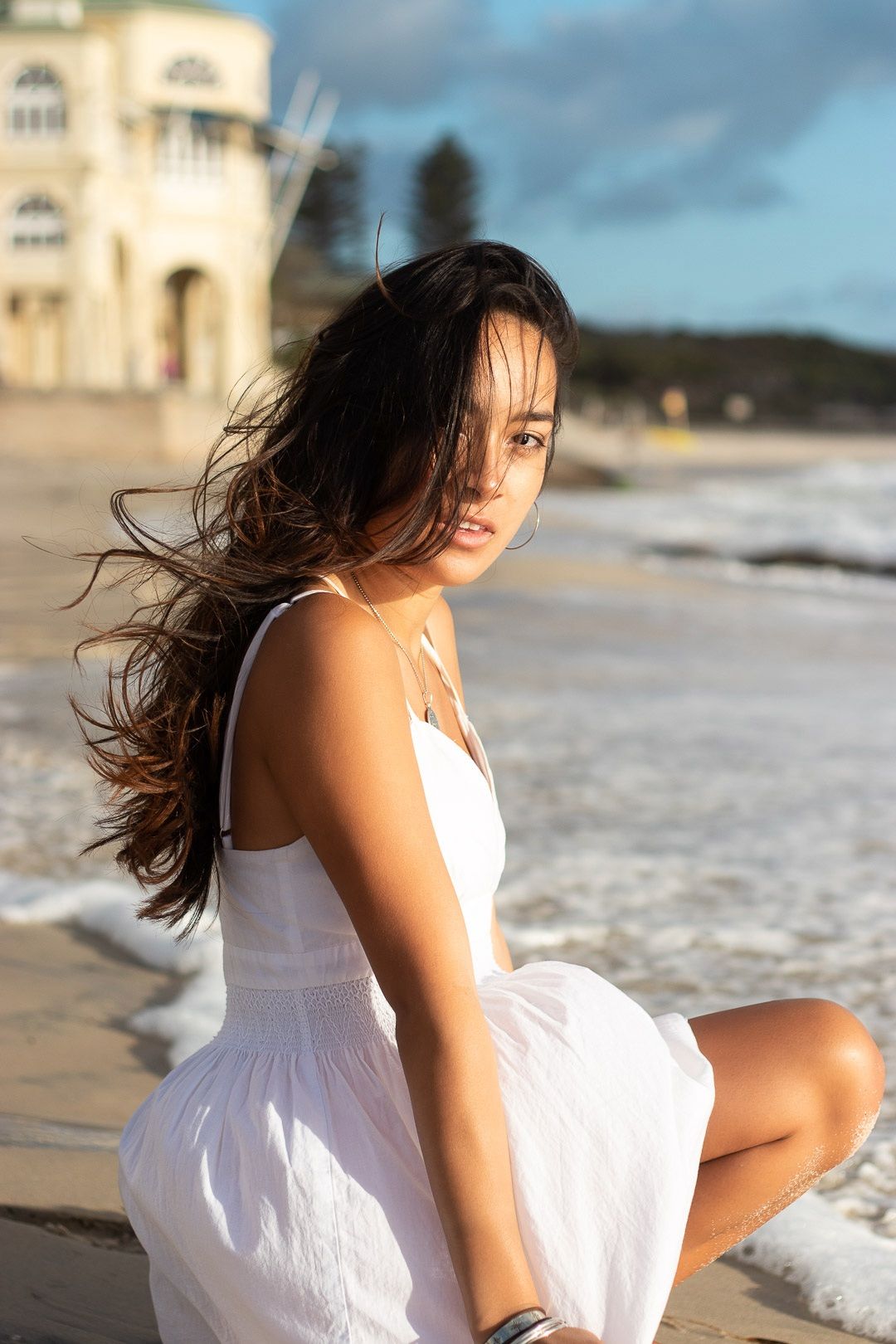 Model in flowing white dress poses on beach at sunset with waves in background.