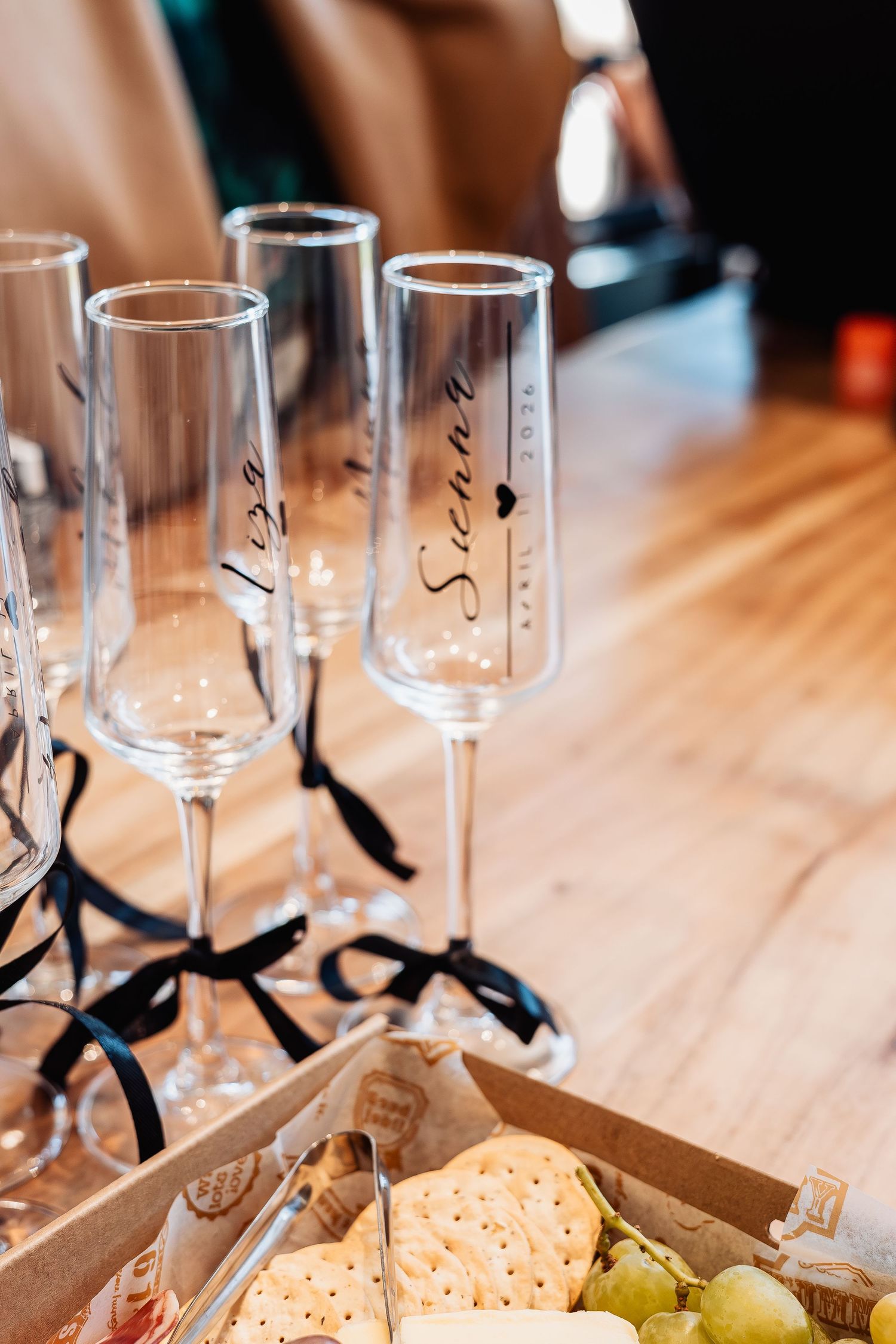 Personalized champagne flutes with black ribbon bows and calligraphy names displayed on wooden table with charcuterie board.