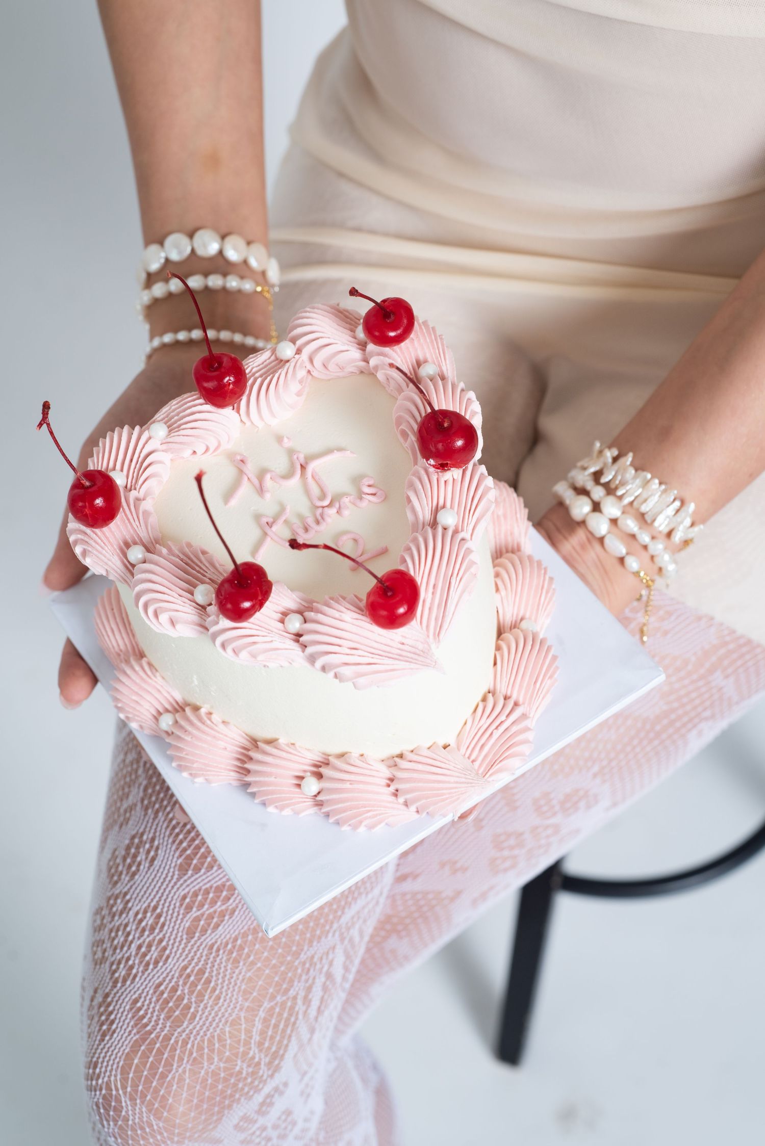 Close-up of hands holding decorated white cake with pink frosting swirls and cherry garnishes.
