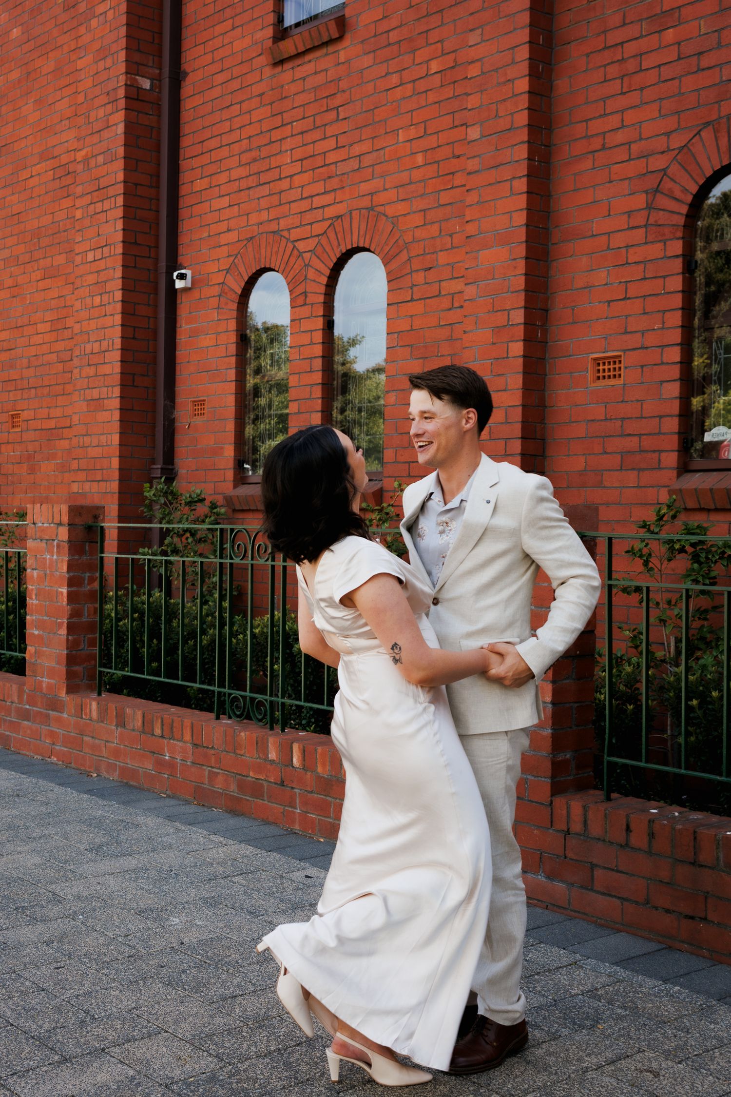 A couple in white outfits sharing a joyful moment against a brick wall backdrop.