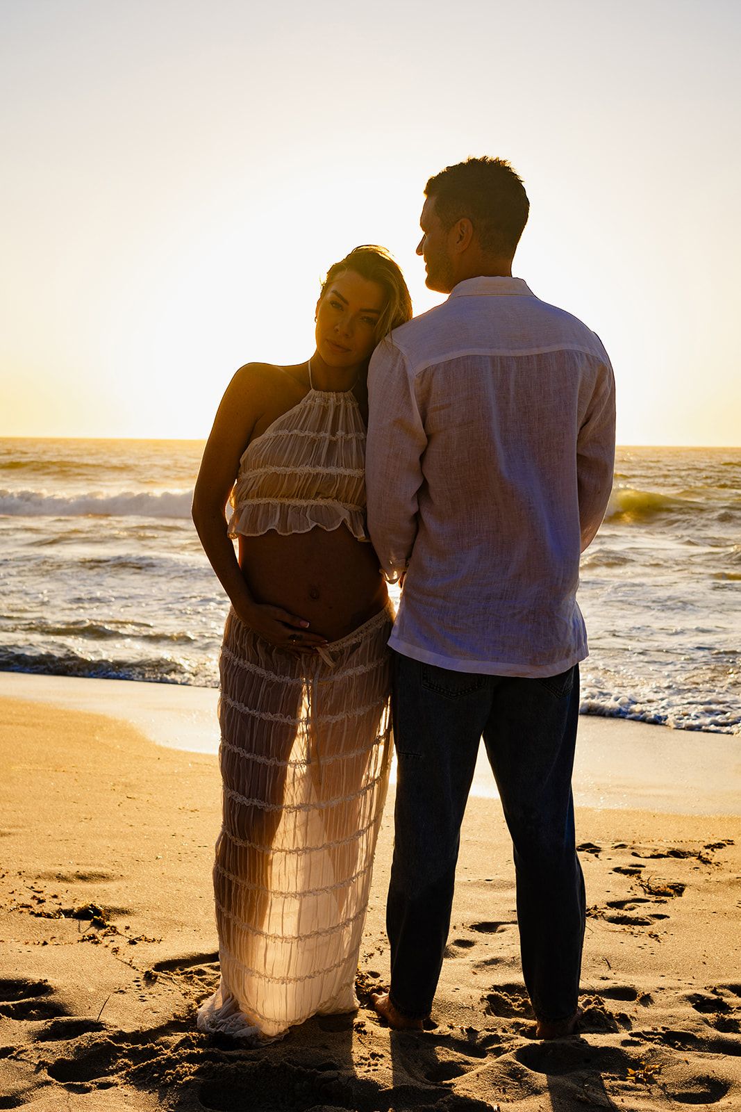 Couple standing together on sandy beach at sunset watching ocean waves.