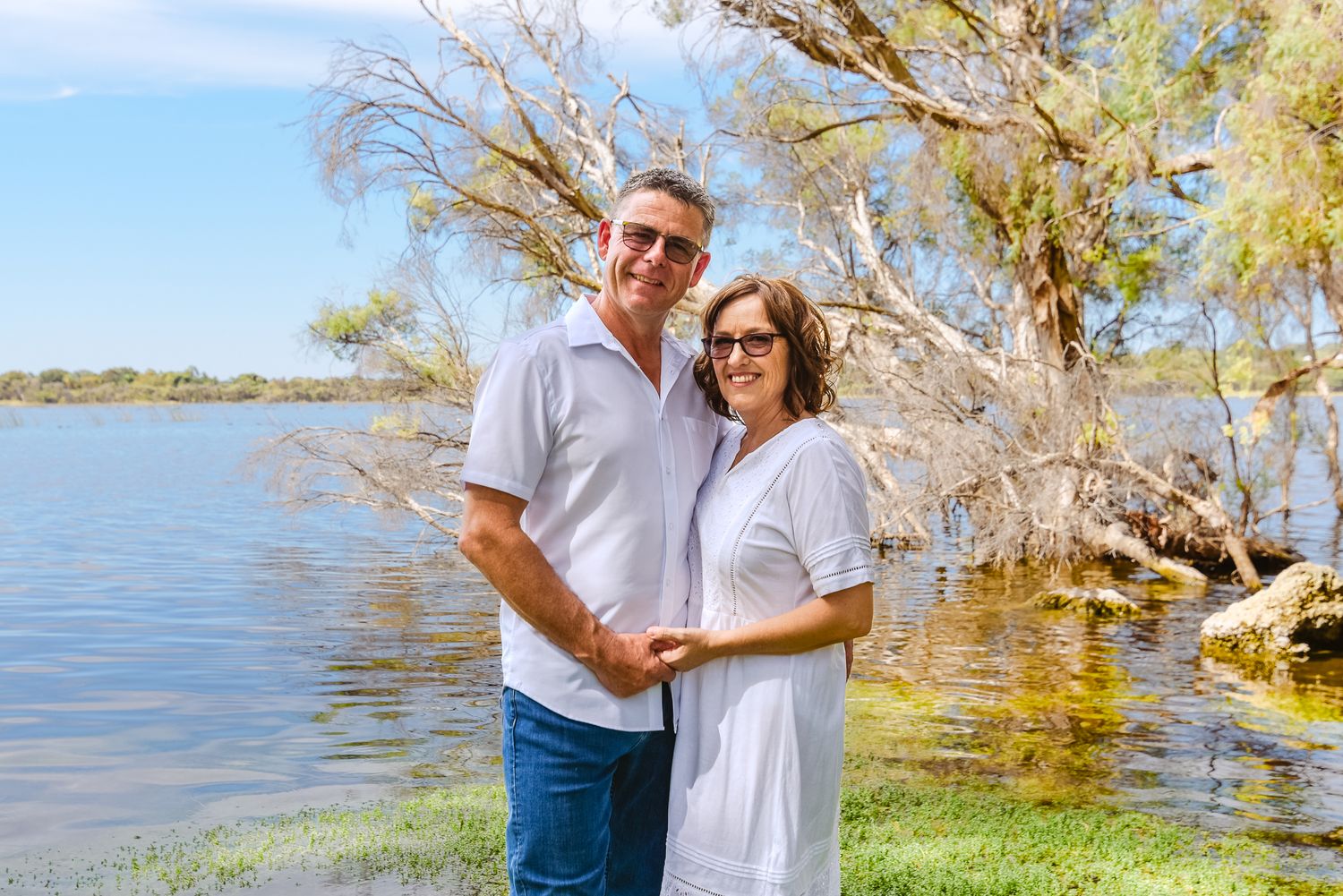 Romantic couple poses along lakeside path with trees and water in scenic background.