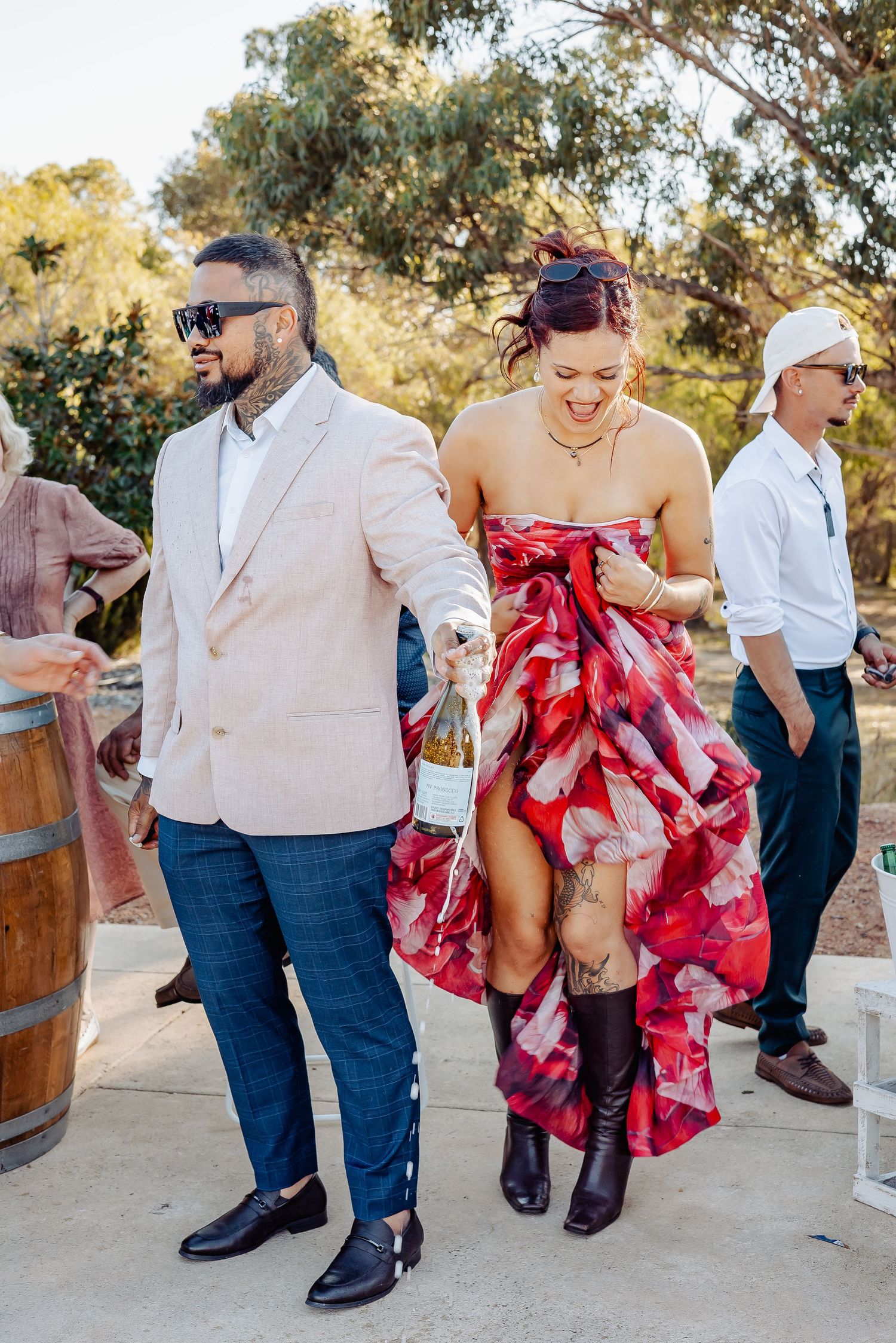 Couple in formal attire holding wine bottle at outdoor wedding, woman in floral dress with cowboy boots, man in beige blazer.