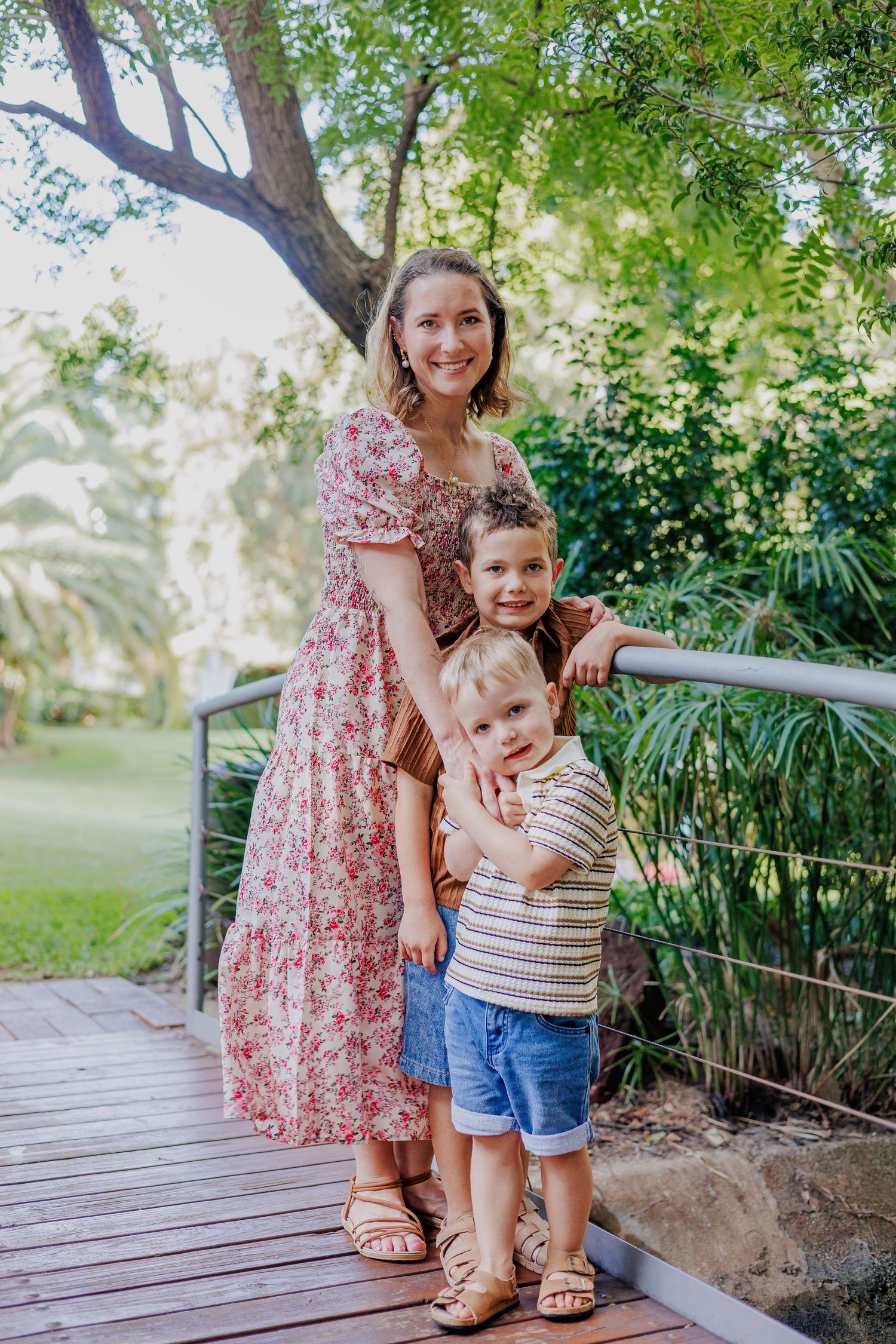 A smiling woman in a floral dress poses with two young boys on a wooden bridge surrounded by lush green trees.