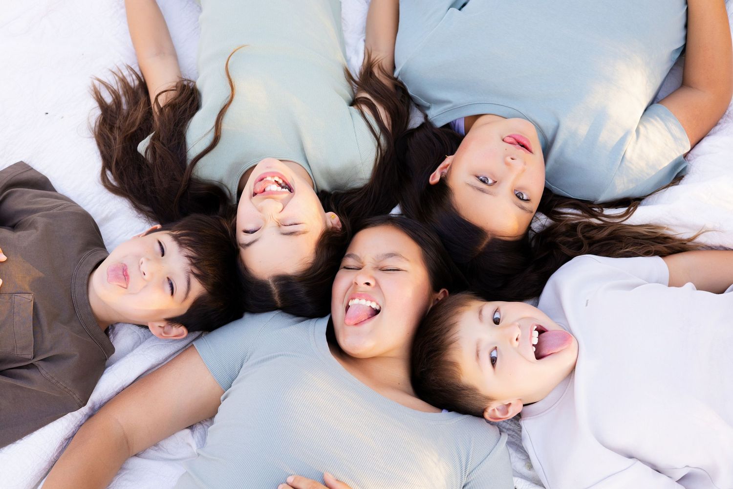 Five children lying in a circle on the ground, smiling and sticking out their tongues in a playful overhead photo.