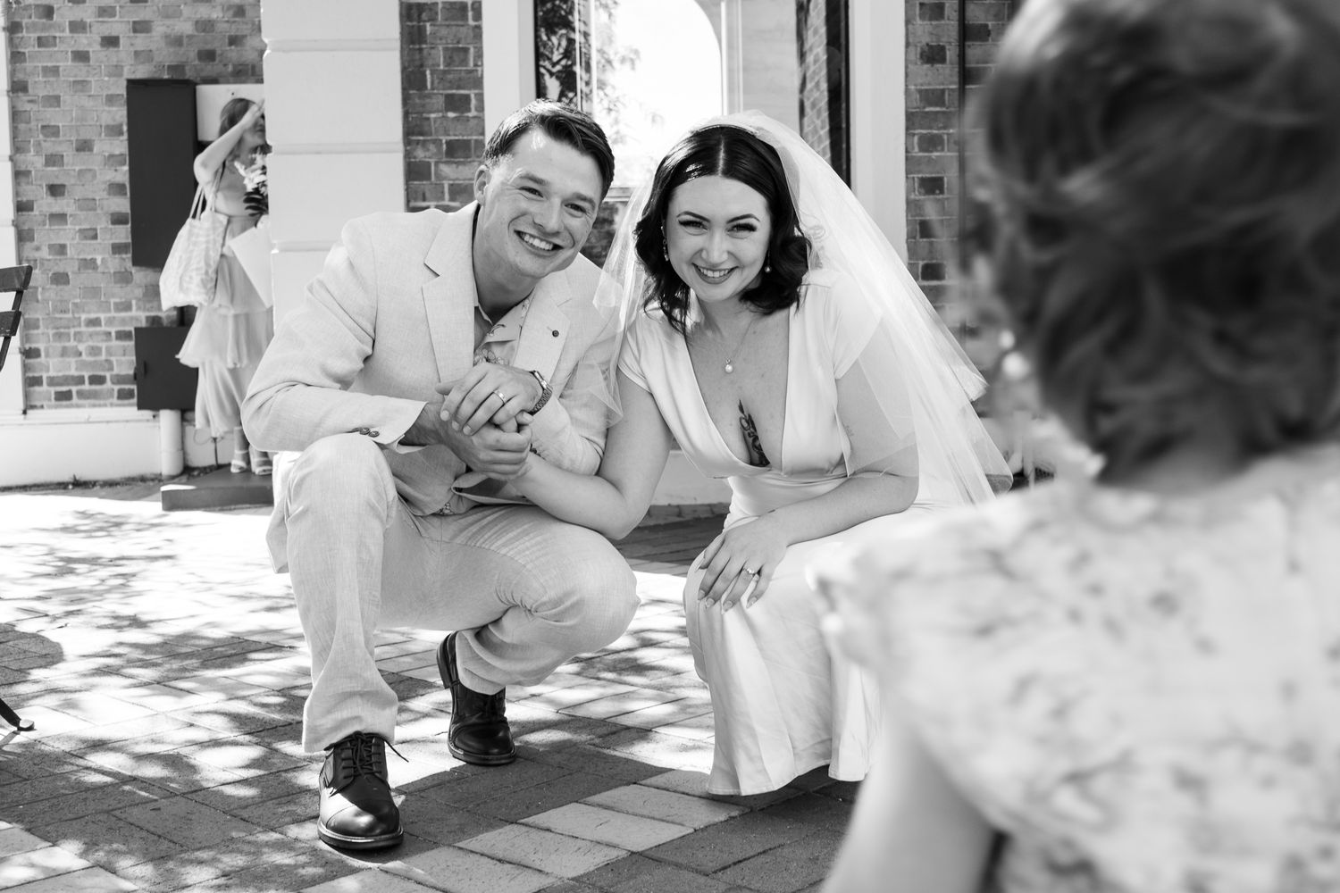 Black and white photo of a smiling bride and groom crouching down together at their outdoor wedding ceremony.