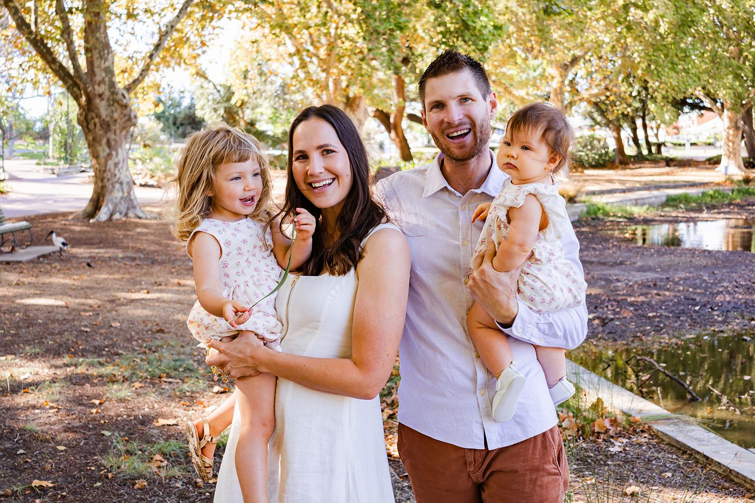Family photoshoot with parents and children in matching white outfits under trees in a park setting.