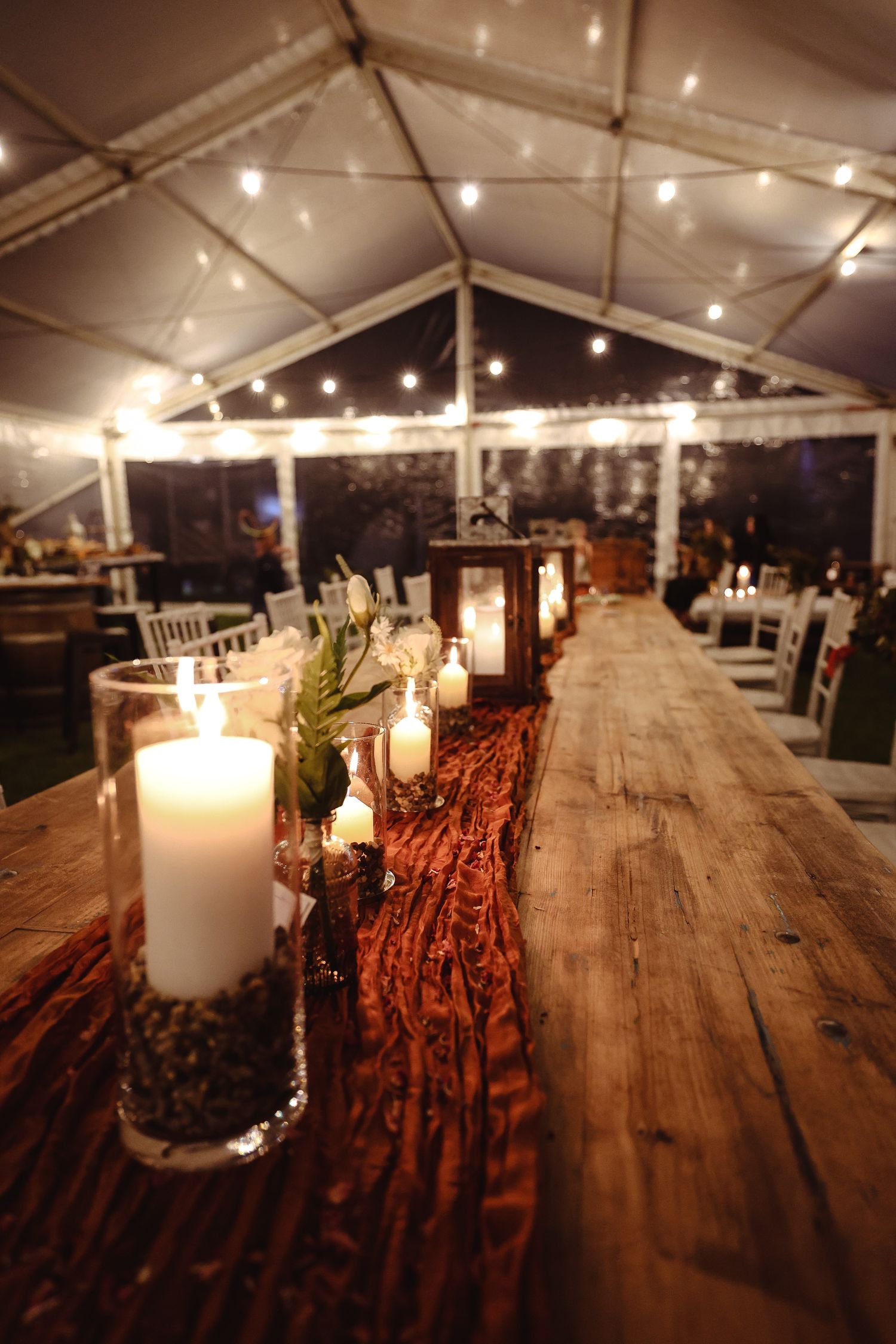 Rustic wedding reception tables with wooden surfaces and string lights under a white tent canopy.