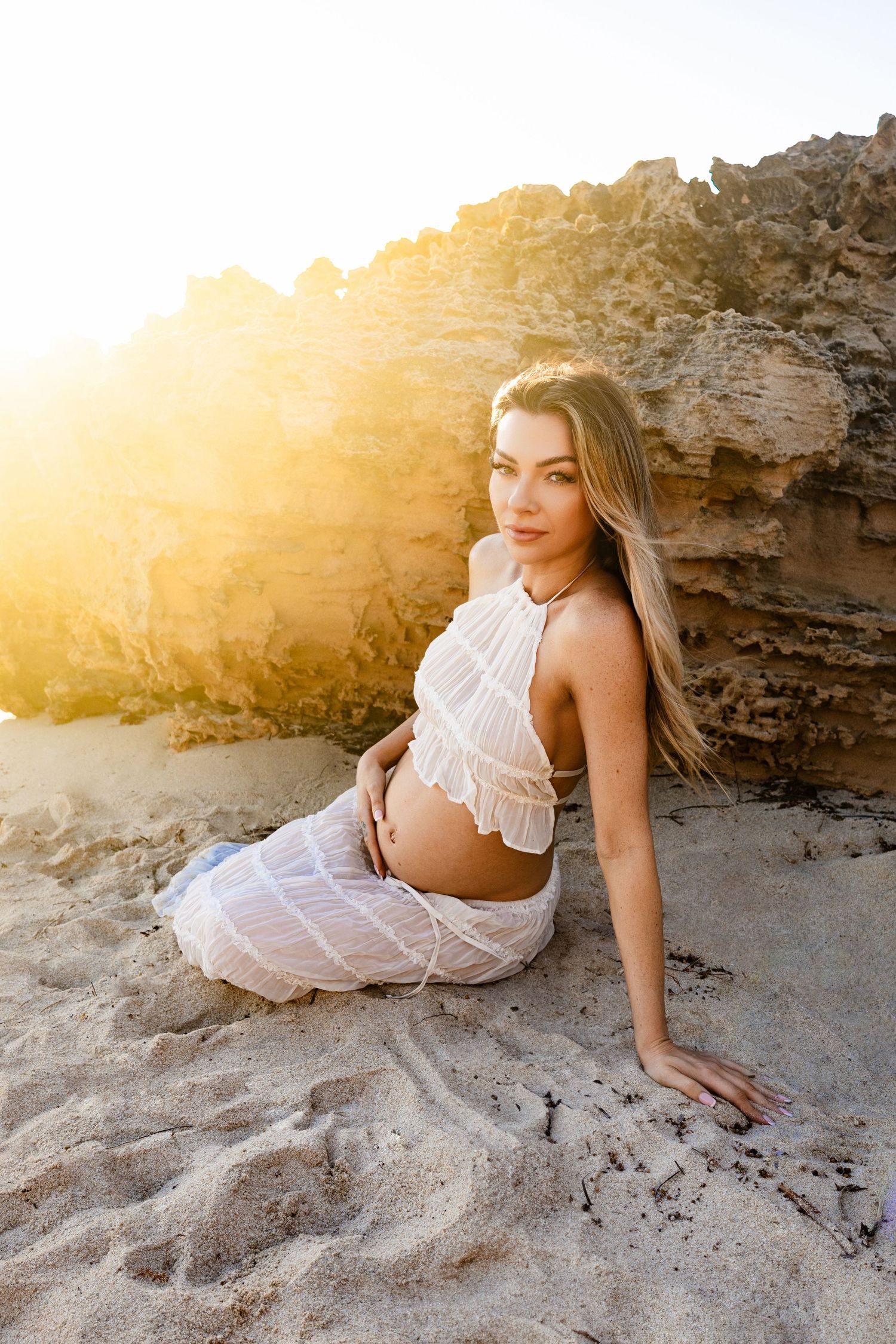 A person in a white dress poses gracefully on rocky terrain during golden hour sunset.