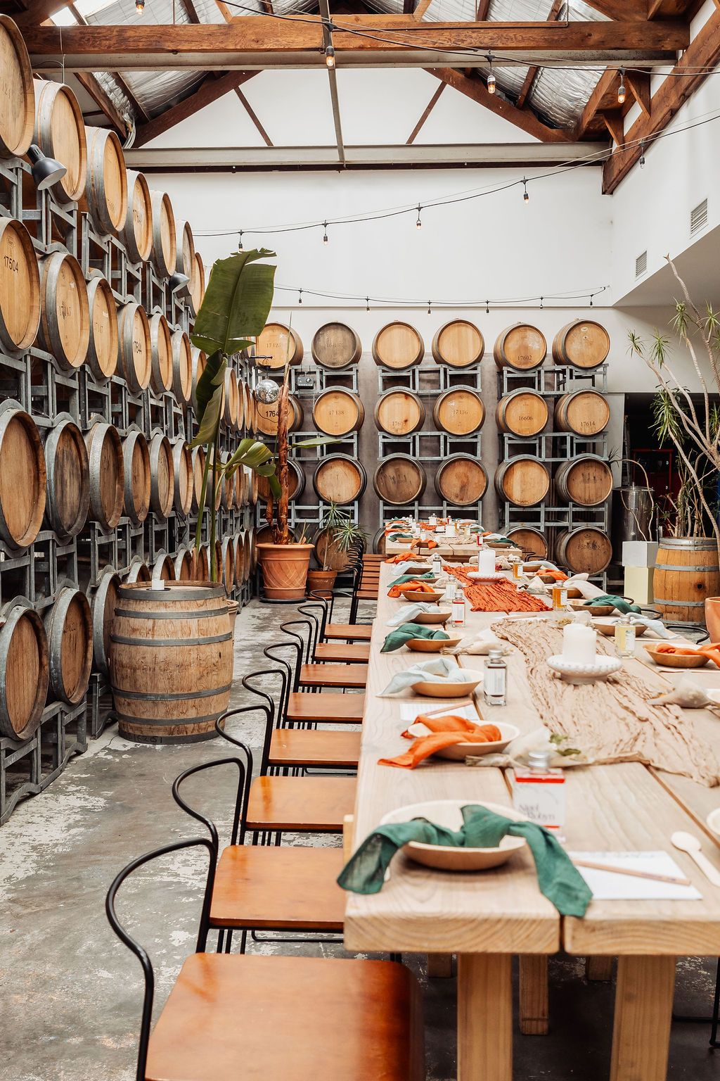 Rustic winery dining room with long wooden tables set for a feast, surrounded by oak barrel walls and string lights.