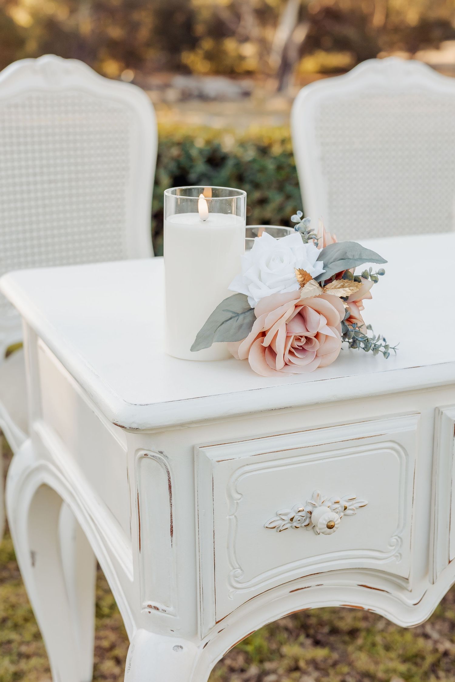 Elegant white vintage table with lit candle and blush pink roses, styled for an outdoor wedding ceremony.