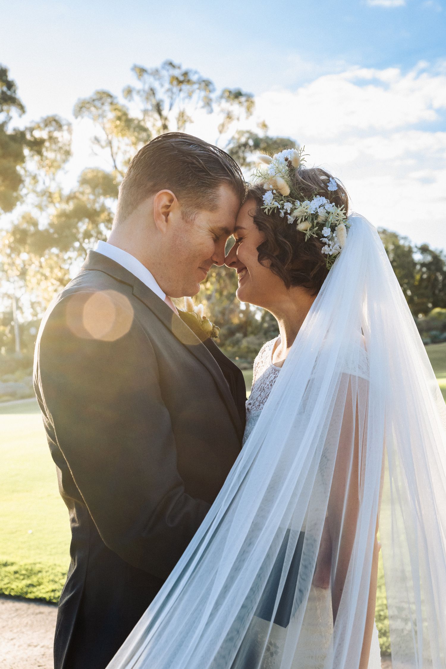 Bride and groom touching foreheads romantically outdoors, bride wearing floral crown and veil in golden sunlight.