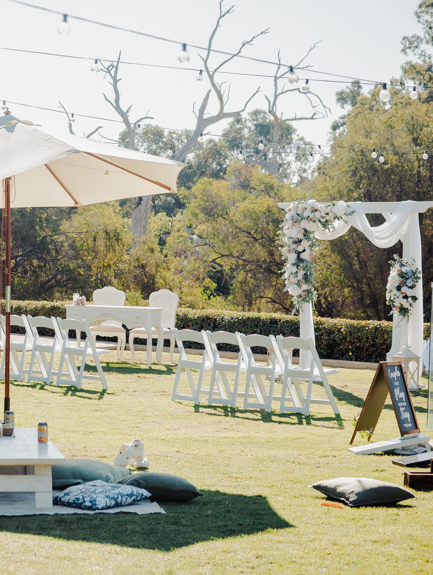 Outdoor wedding ceremony setup with white folding chairs, floral arch, string lights, and cushioned seating on lush green lawn.