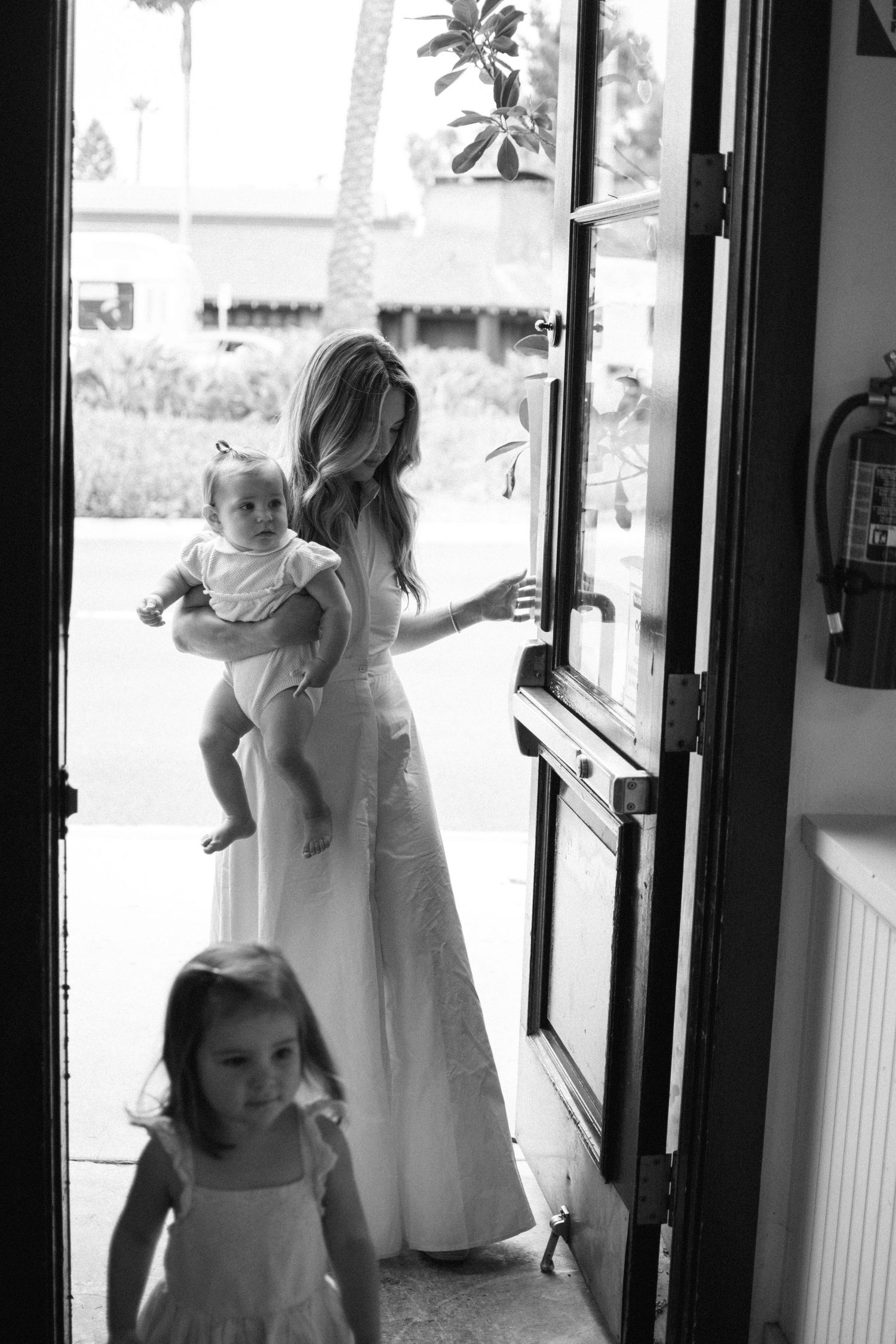 Black and white photo of an elegant mother in a white gown holding a baby while opening a glass door.