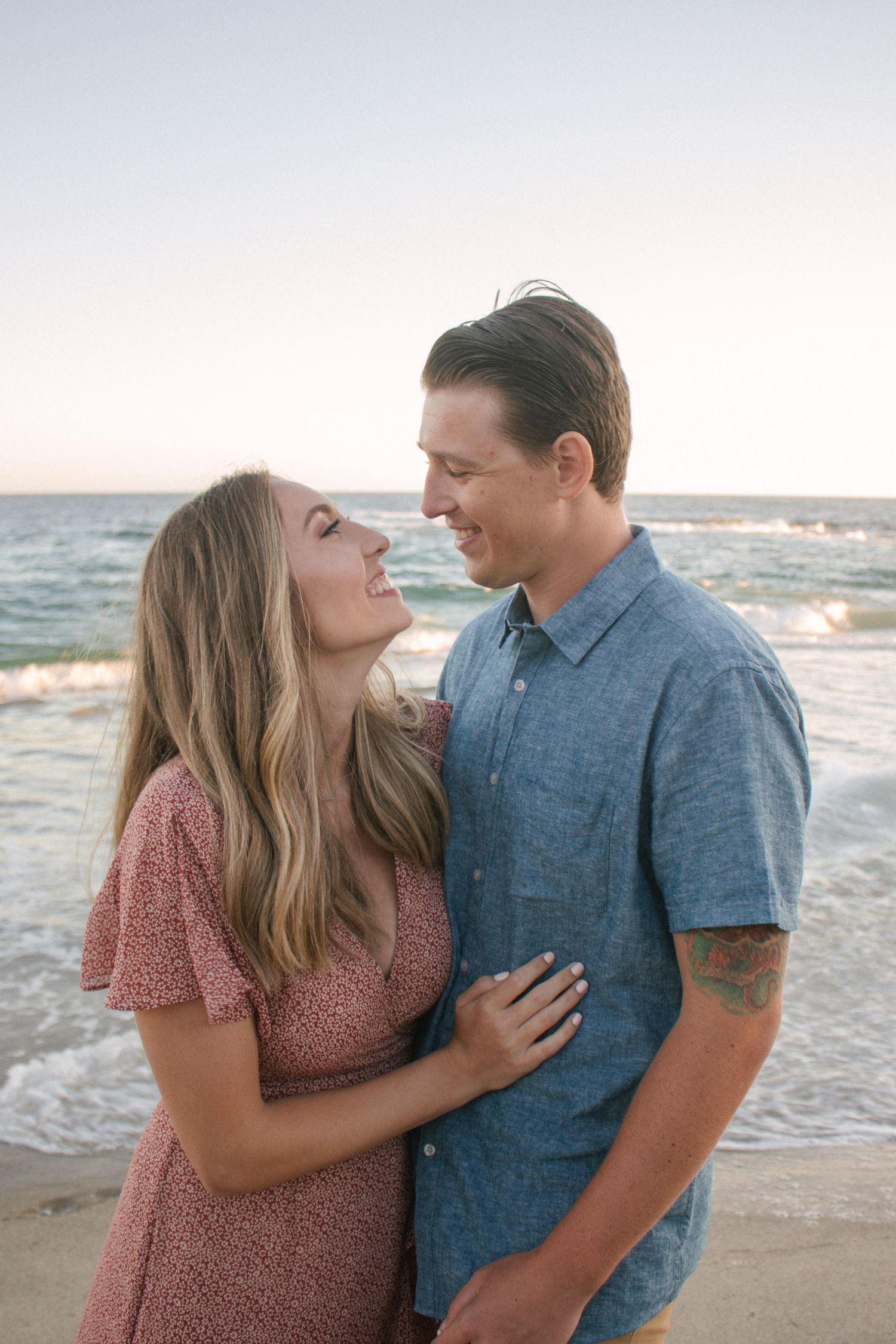 Joyful couple embracing on a beach at sunset, smiling and gazing at each other with ocean waves behind.