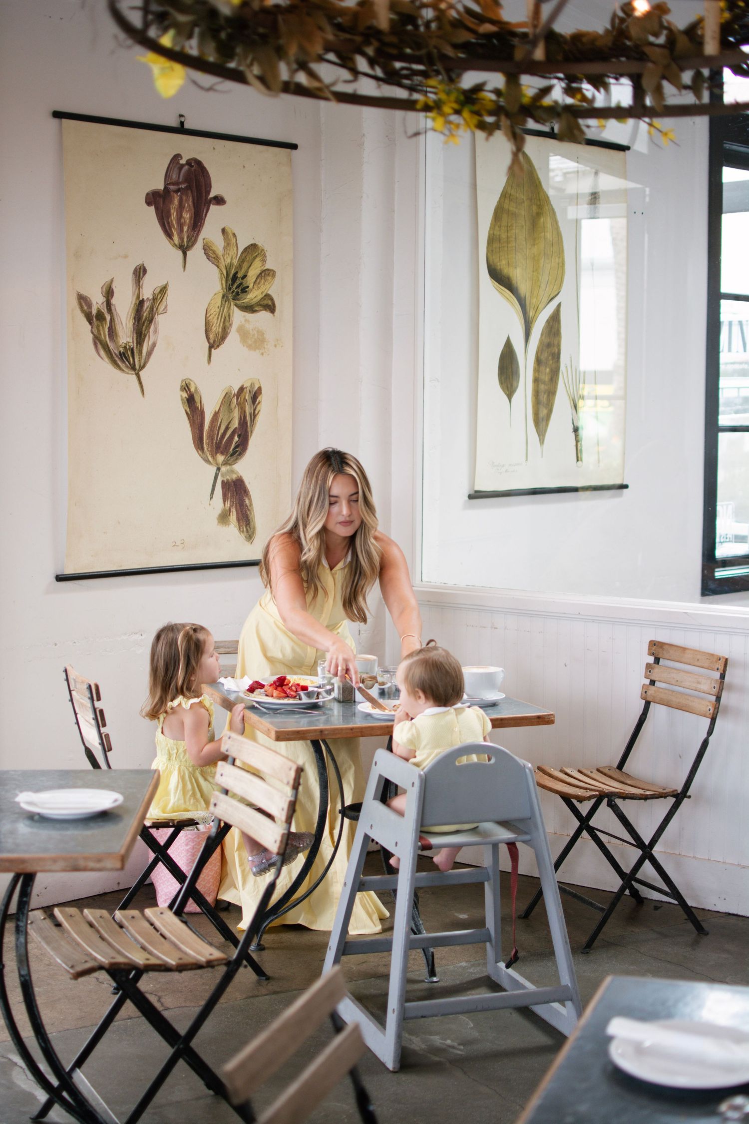 Woman in yellow dress serving food to two young children at a cafe table with botanical artwork on white walls.