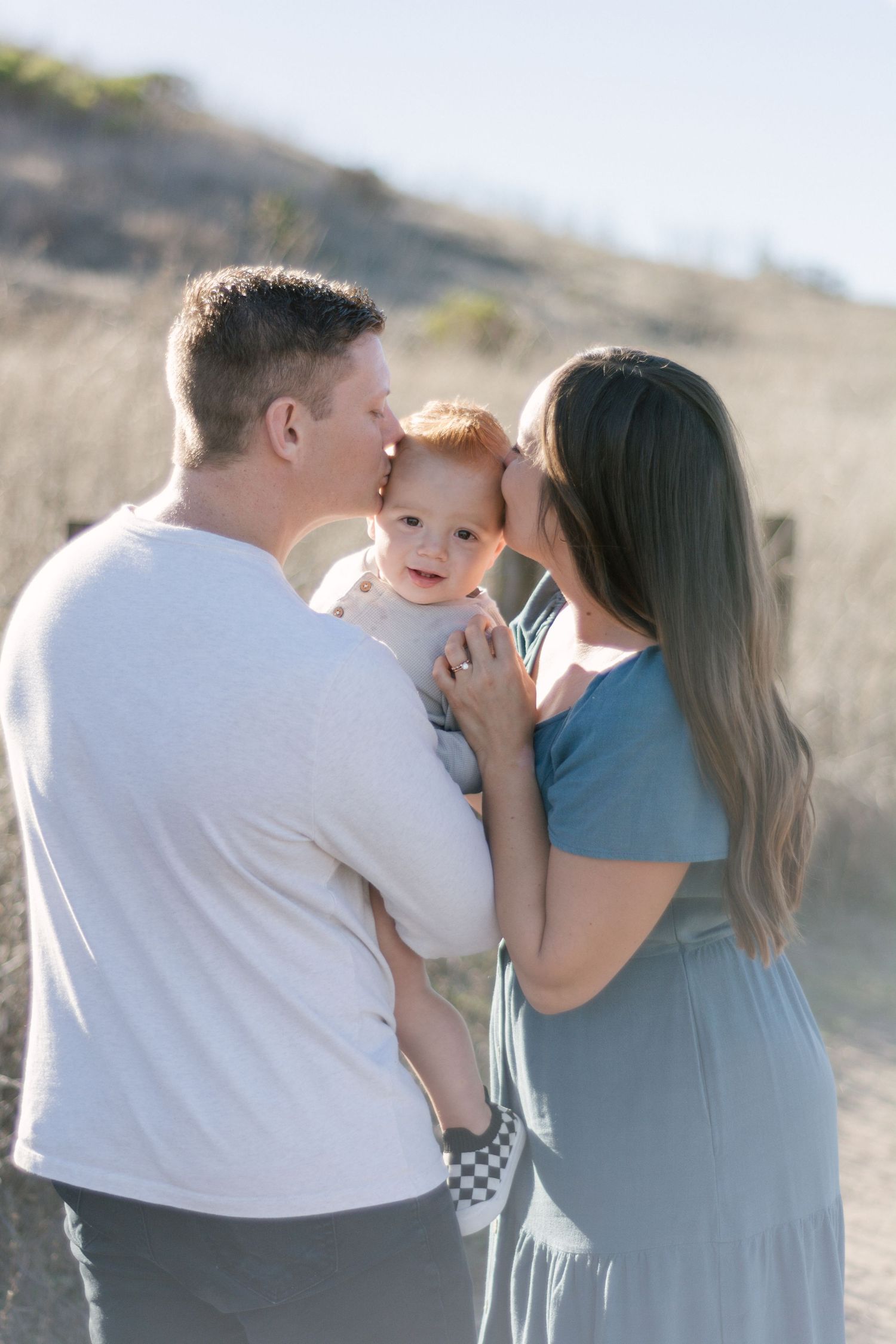 Loving parents kiss their happy red-haired baby on a sunny outdoor dirt trail path.