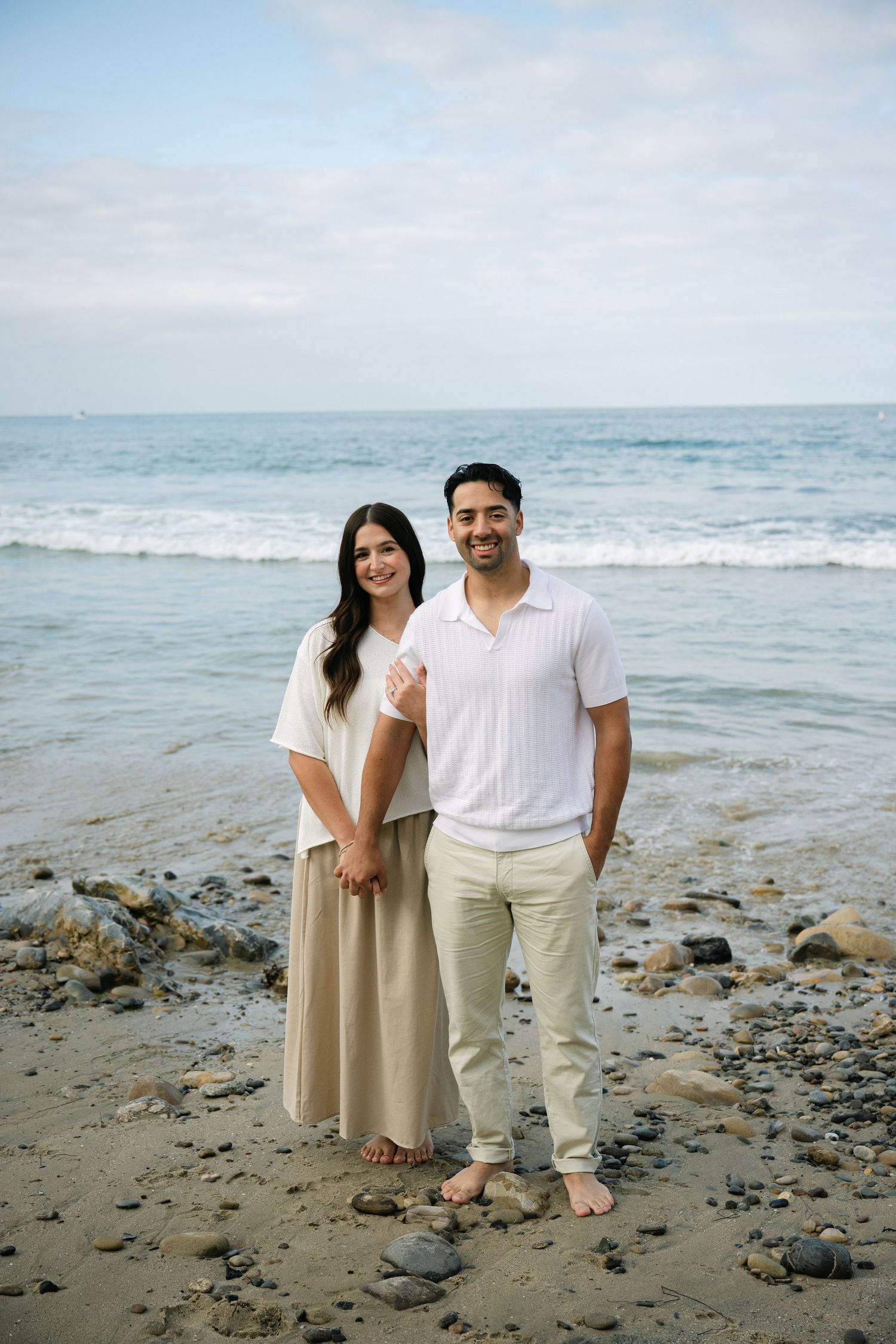 Couple smiling together on a rocky beach with ocean waves in the background.