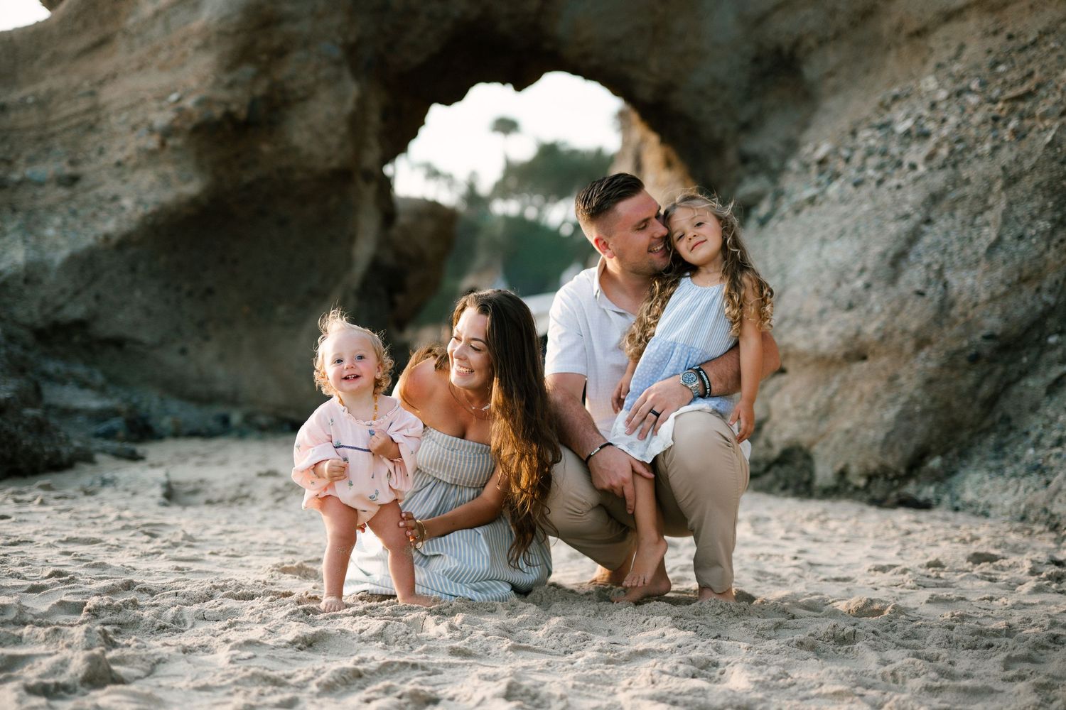 Happy family of four posing together on sandy beach near large rock formation in warm golden light.