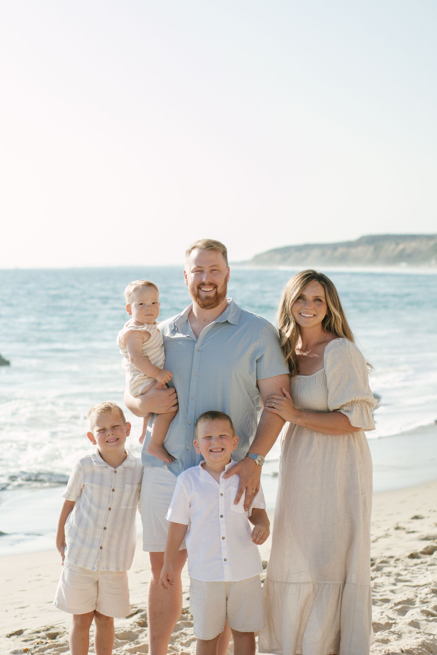 Happy family of five posing on a sunny beach, parents holding children with ocean cliffs behind them.