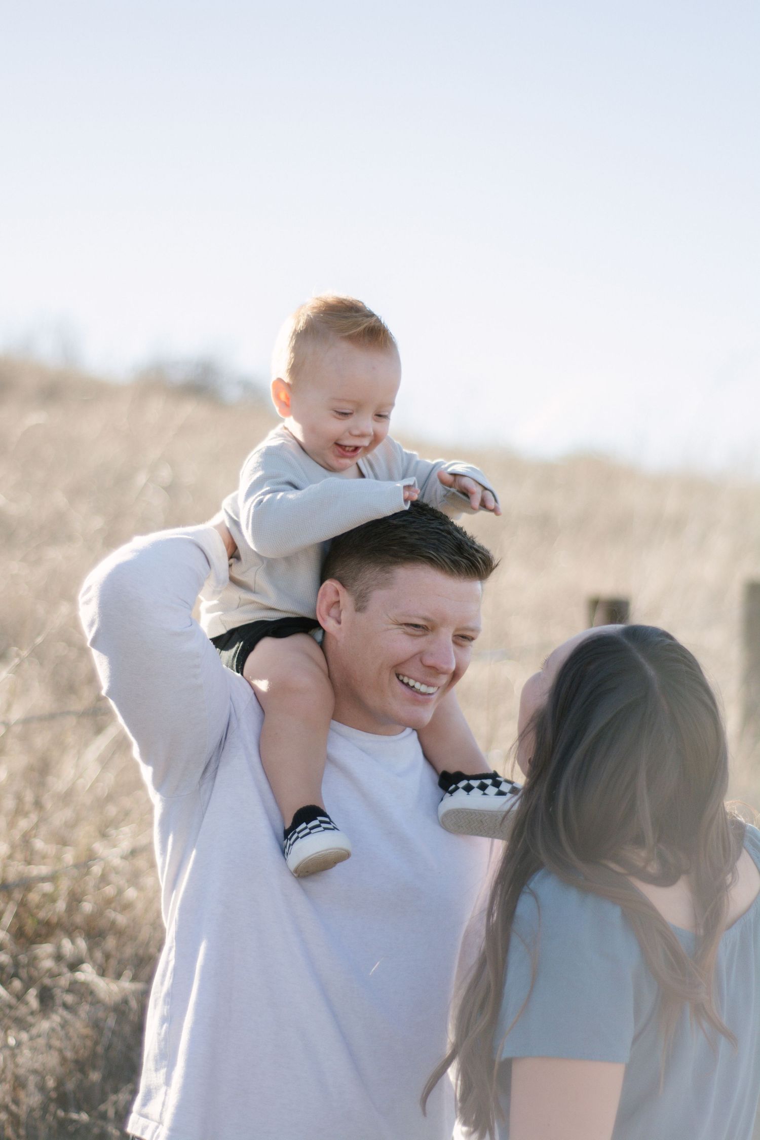 Laughing father carries smiling baby on shoulders while mother looks on in a dry field.