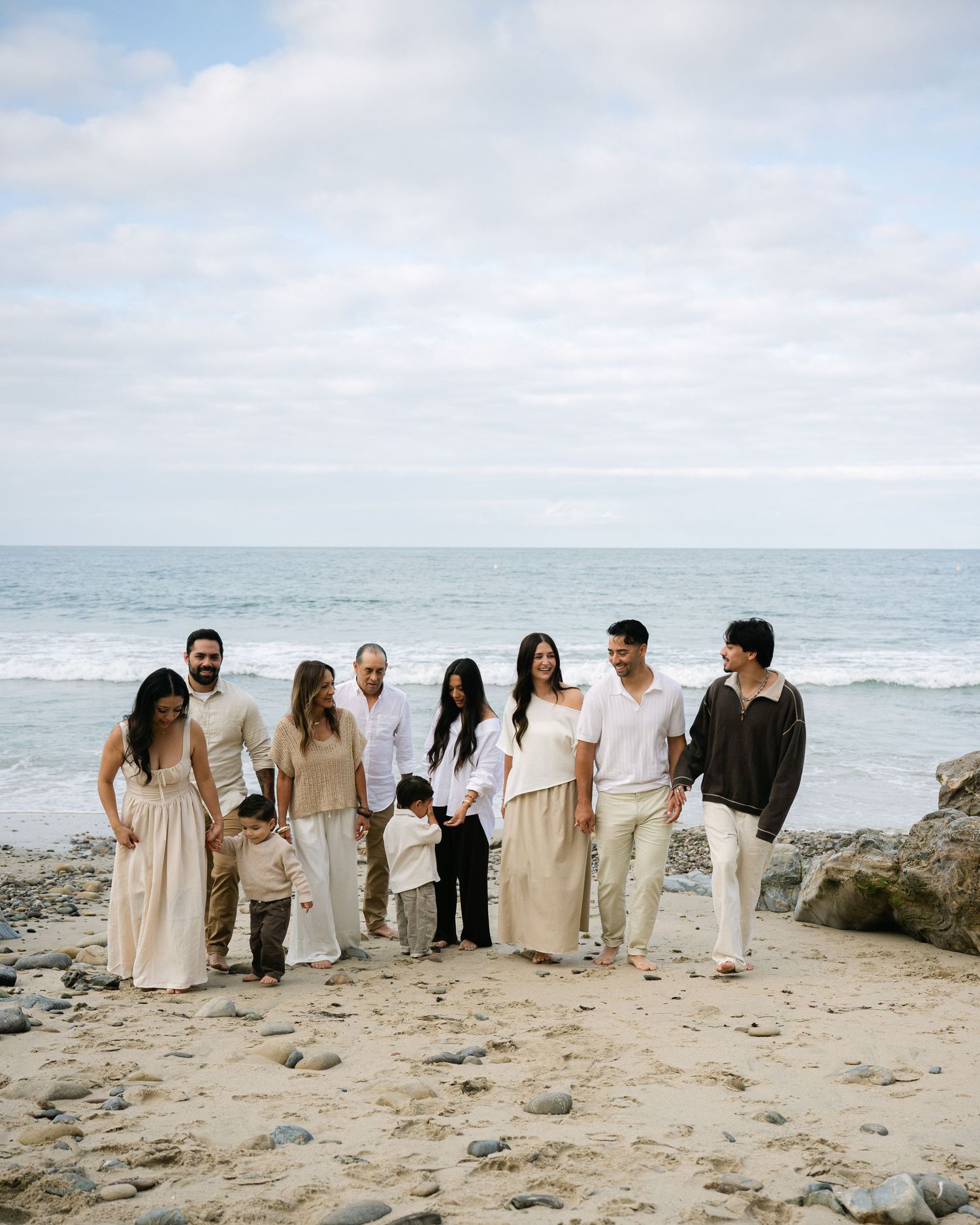 Large family group posing together on a rocky beach with ocean waves and cloudy sky in the background.