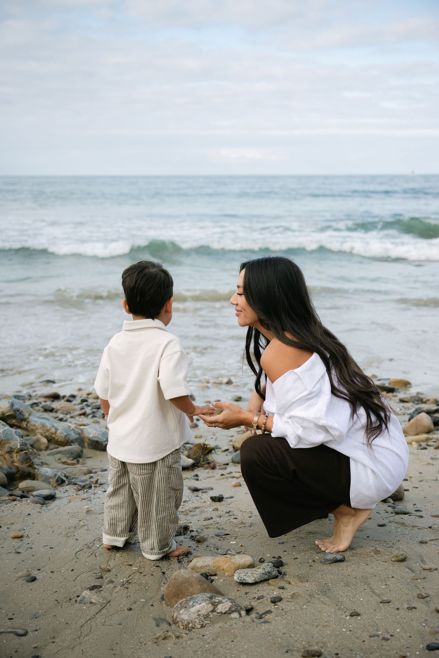 A mother crouches beside her young son on a rocky beach, sharing a tender moment while ocean waves crash behind them.