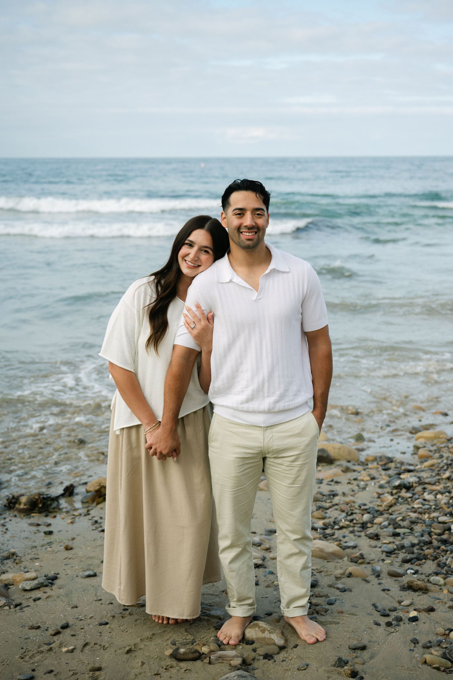 Couple smiling and embracing on a rocky beach shoreline with blue ocean waves and cloudy sky behind them.
