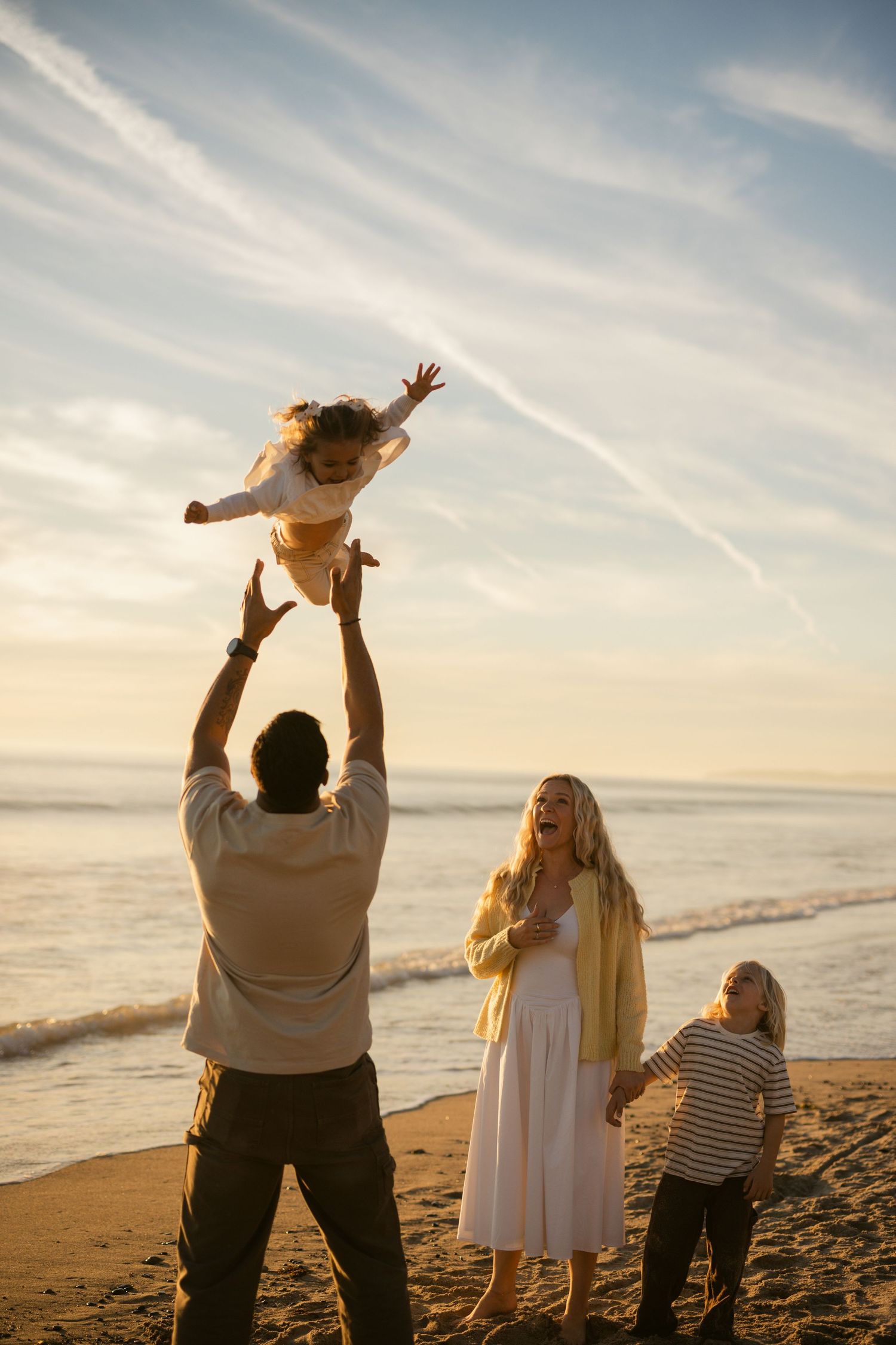 Father joyfully tosses child in the air on a golden sunset beach with family nearby.