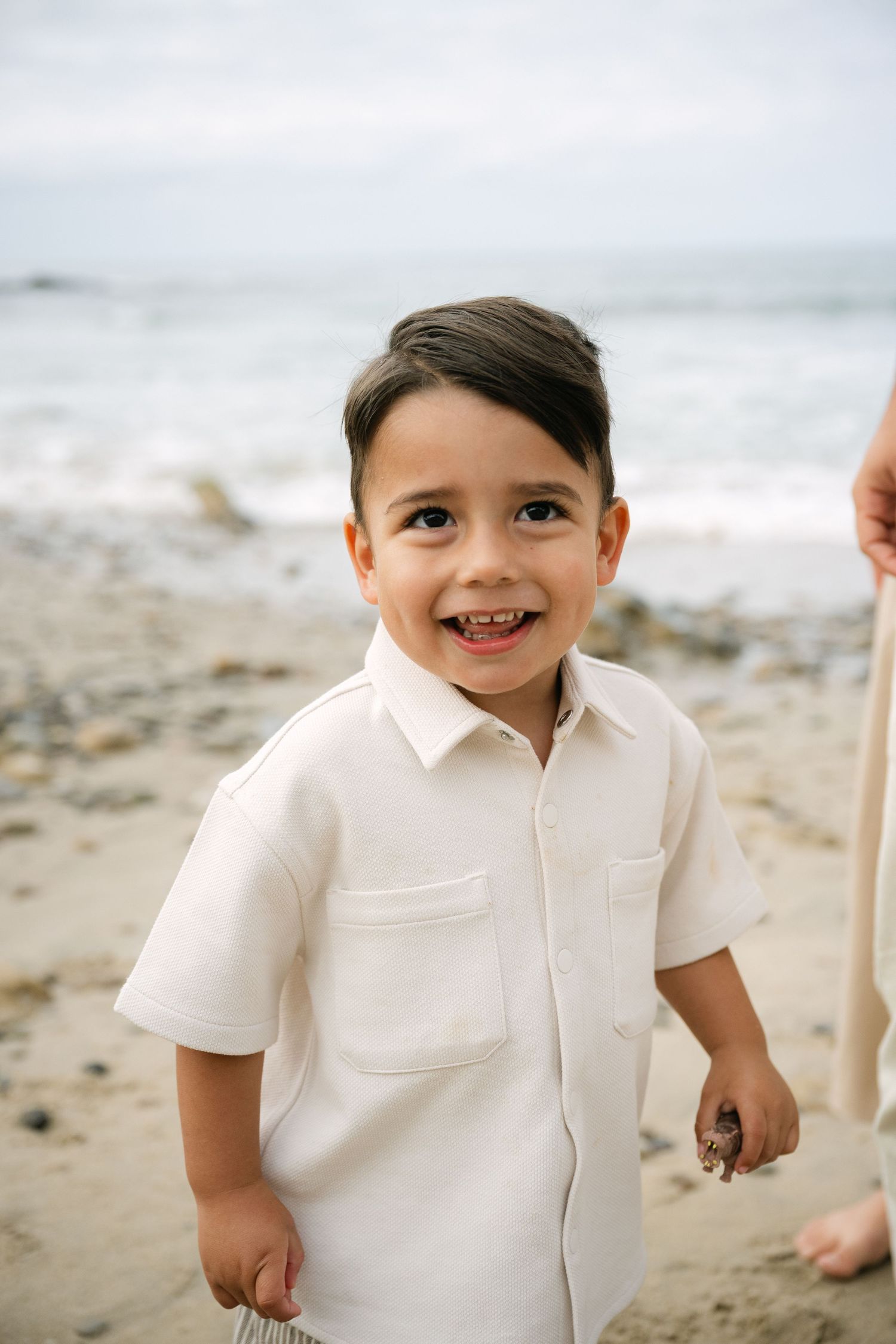 Happy young boy in white shirt smiling at camera on sandy beach with ocean waves behind him.