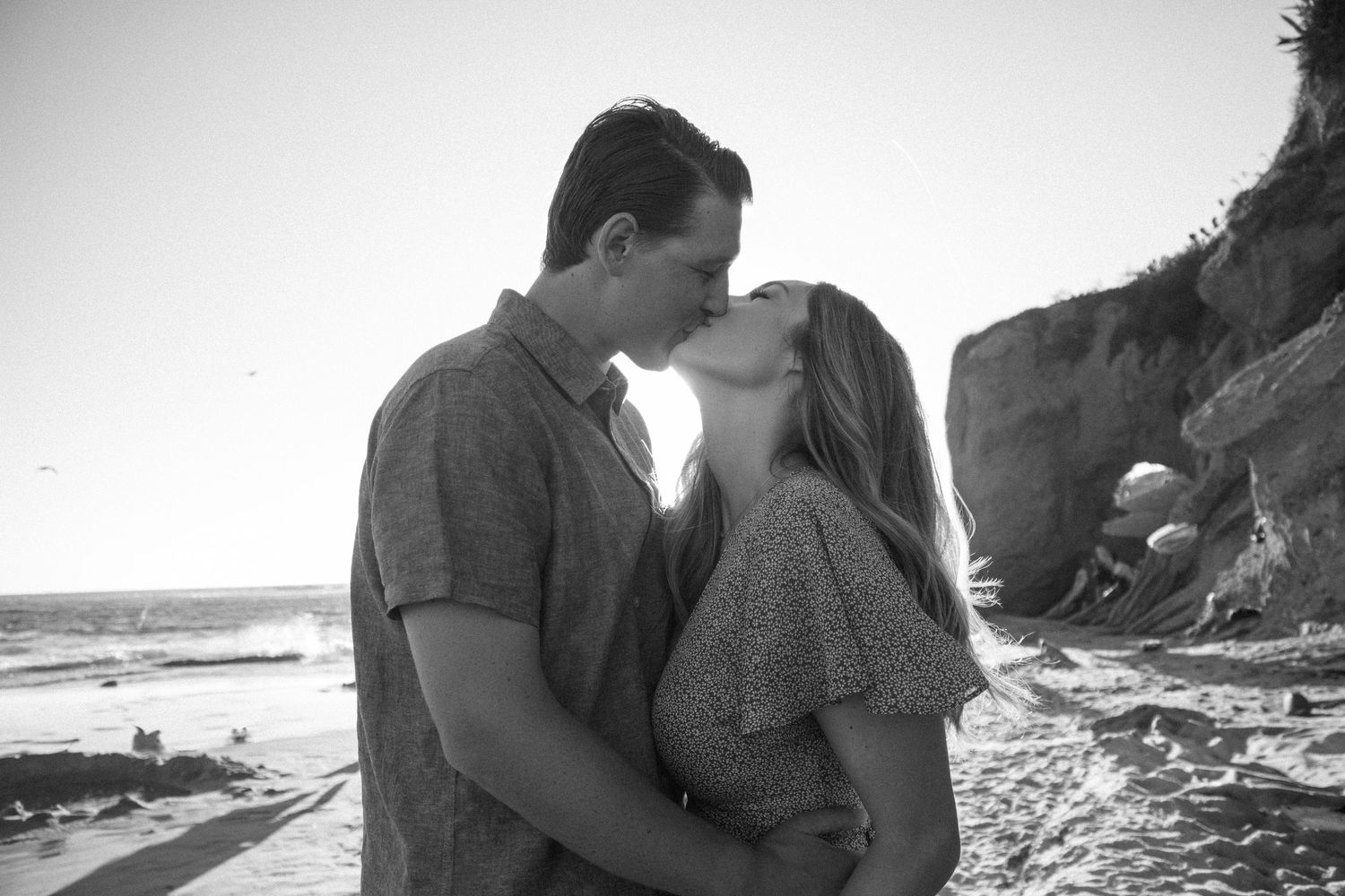Romantic black and white image of a couple kissing on a scenic beach with cliffs in the background.