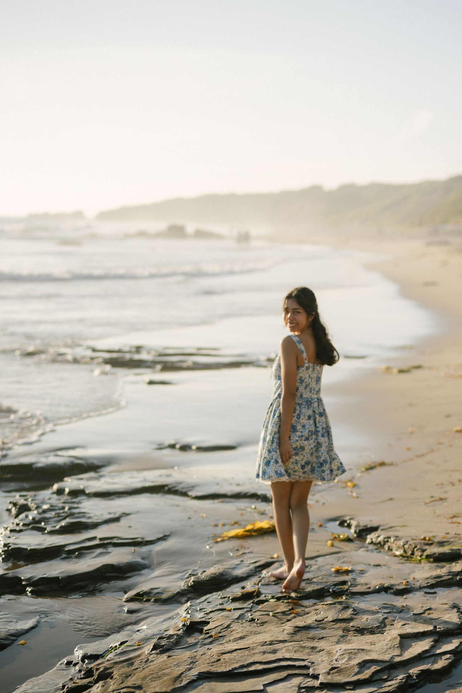 Young woman in a floral dress standing on rocky shoreline at sunset.