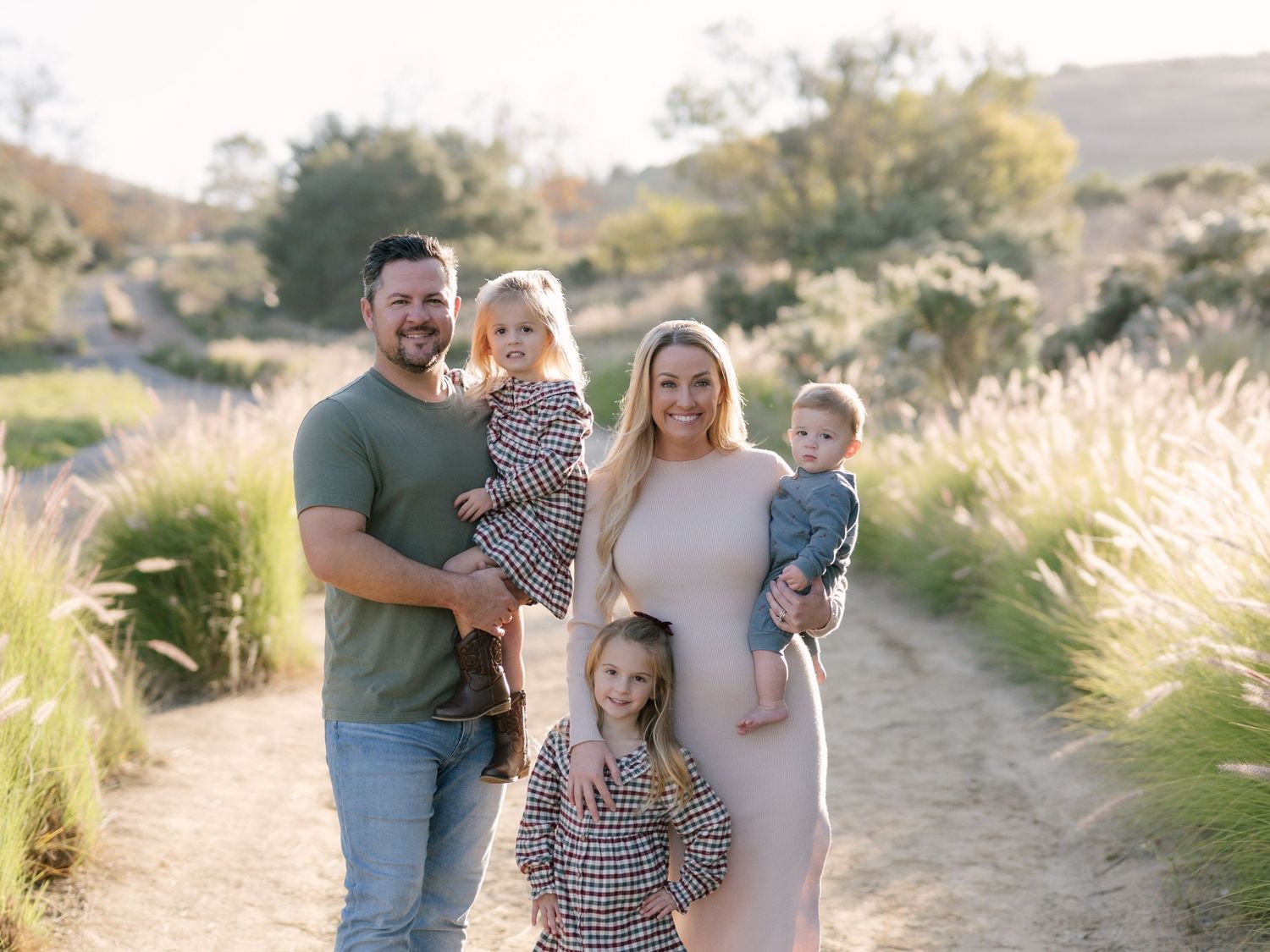 Smiling family of five posing on a sun-drenched nature trail with tall grasses around them.