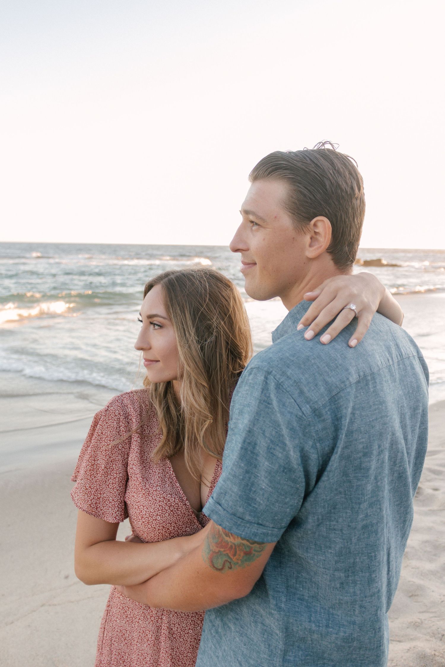 Couple embracing on a beach at sunset, woman resting her hand on mans shoulder as they gaze at the sea.