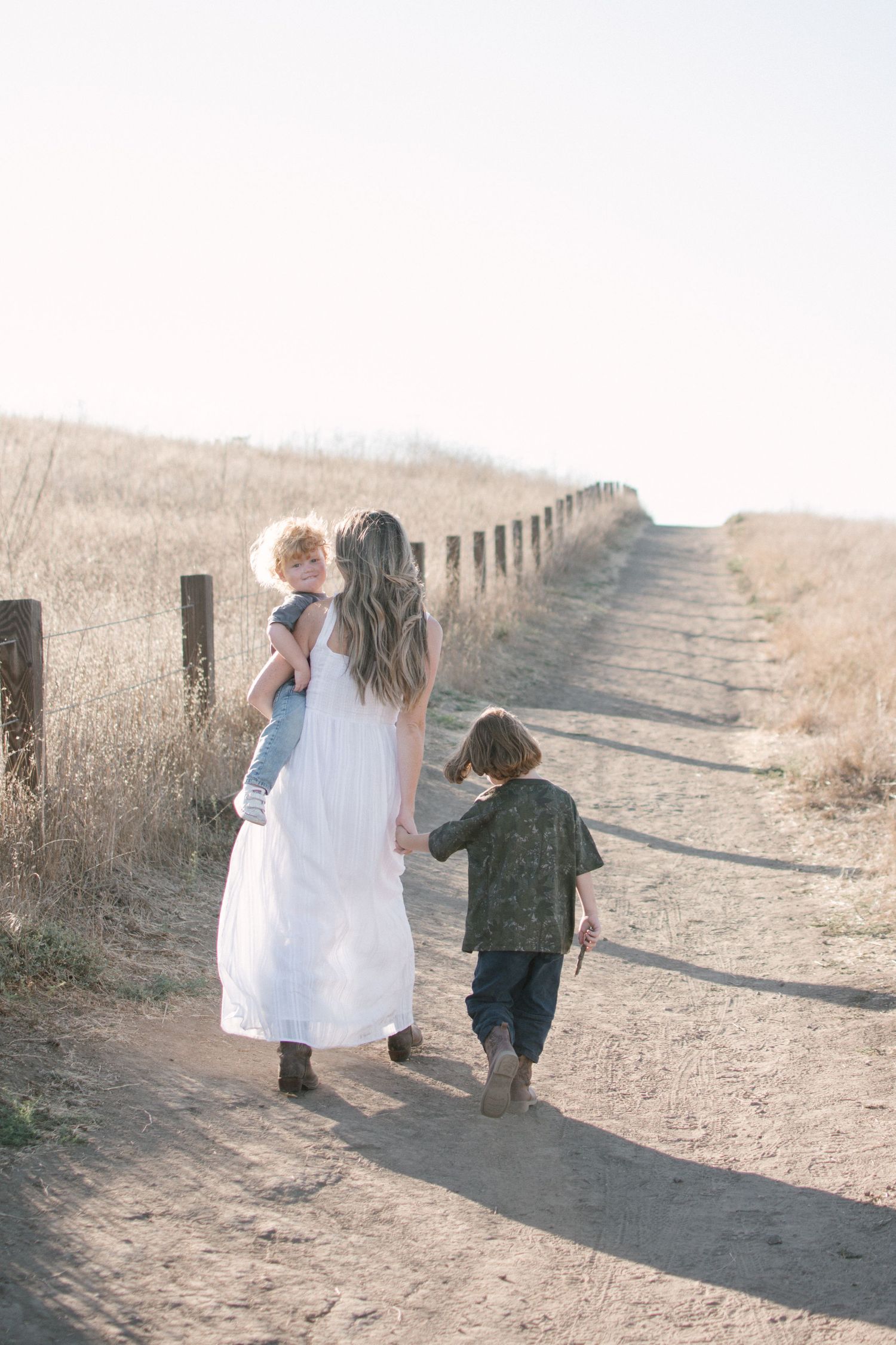 A mother in a white dress walks down a sunny dirt path carrying a toddler while her young son follows.